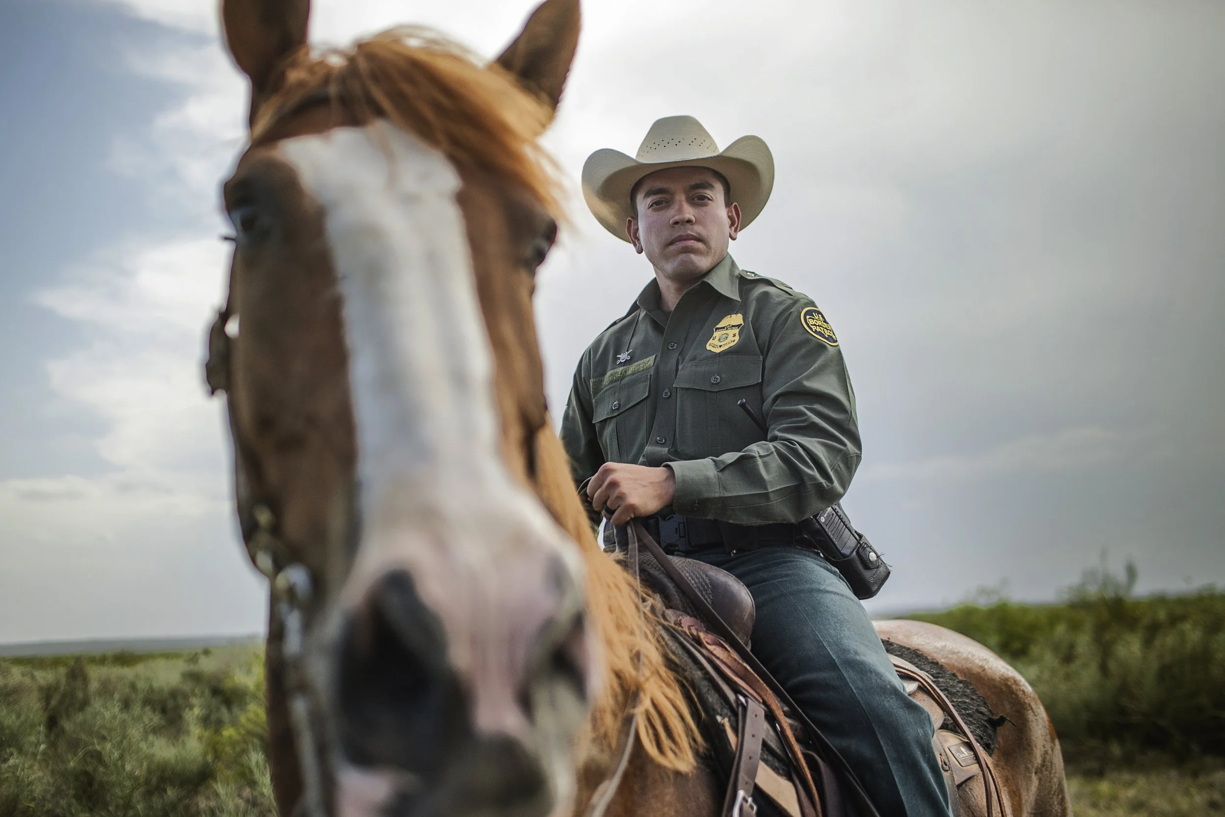 A police officer wearing a cowboy hat sitting on a horse, with a camera attached to his waist, in an outdoor rural setting under cloudy skies.