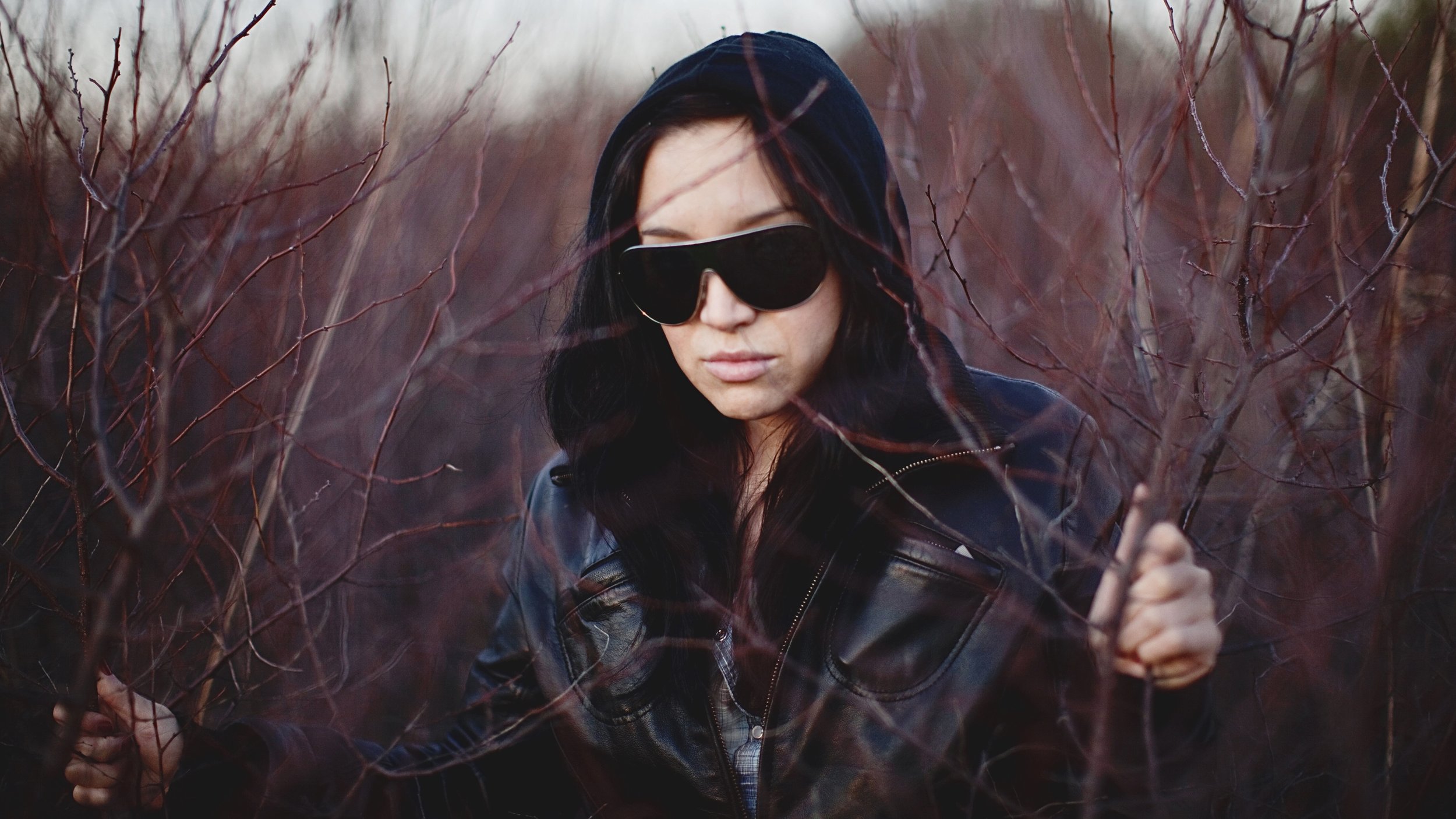 A woman with dark hair wearing black sunglasses and a black hoodie, holding onto tree branches in a thicket of reddish-brown, leafless bushes.