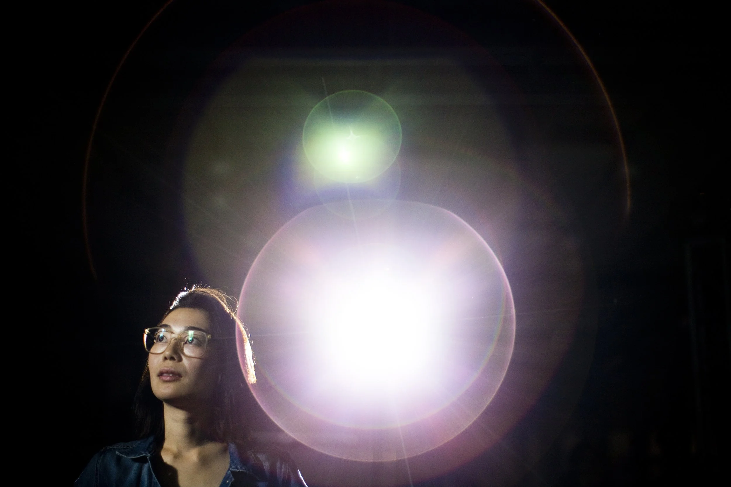 A woman with glasses looking to the left, illuminated by bright, colorful lens flare and light effects in a dark environment.