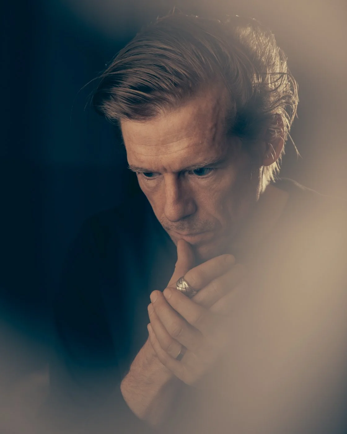 A man with light brown hair looking down thoughtfully, his hand resting on his chin, wearing rings on his fingers.