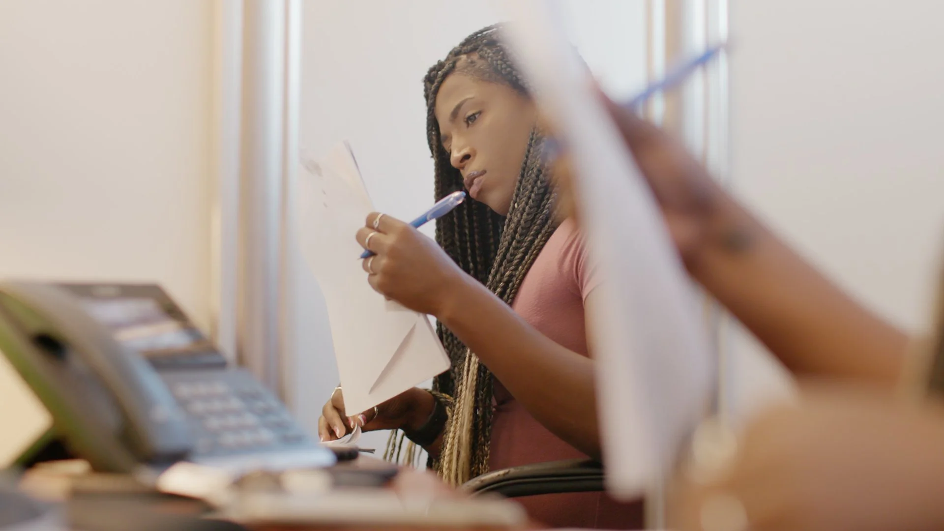 A woman with braided hair looks at a document while talking on the phone in an office or workspace.