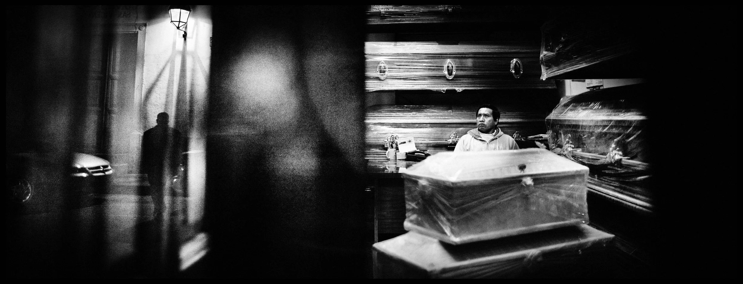 A black and white photo of a man sitting behind a counter in a store with shelves filled with boxed products wrapped in plastic, while a blurred silhouette of a person walking outside can be seen through a window in the foreground.
