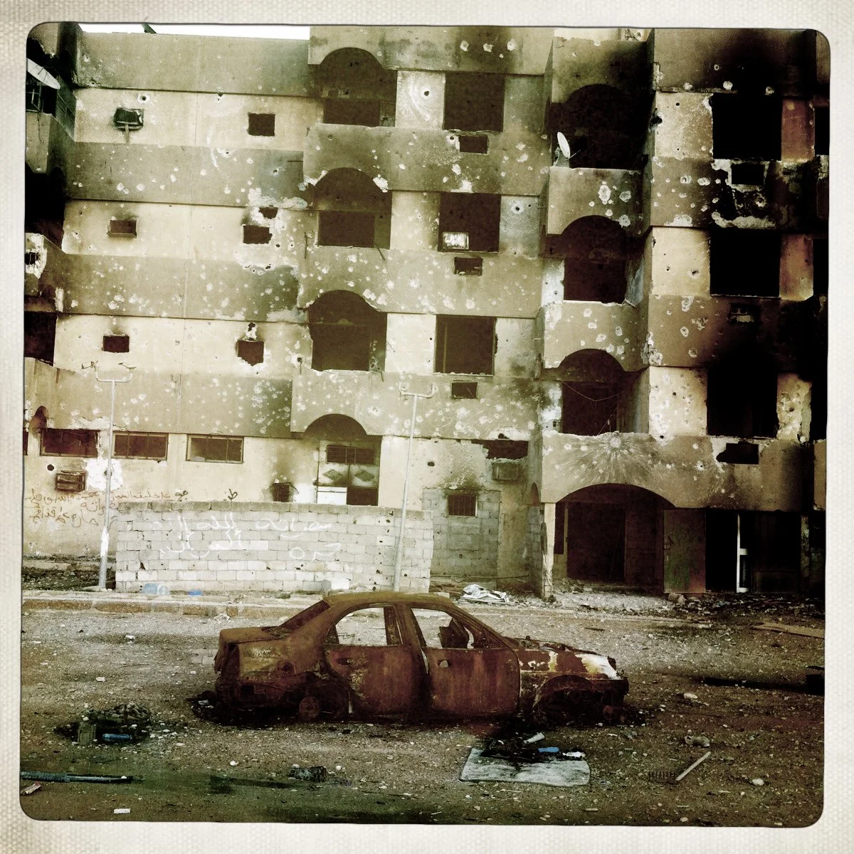 A burnt-out, rusted car in front of a damaged, bullet-riddled building with multiple empty window openings.