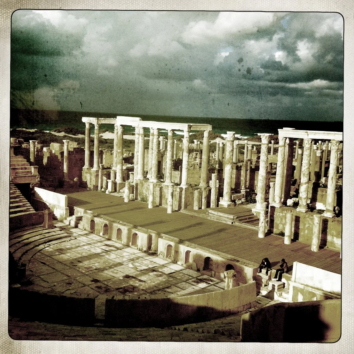 Ancient Roman theater with stone columns and ruins, dark cloudy sky in the background, and two people sitting on the steps.