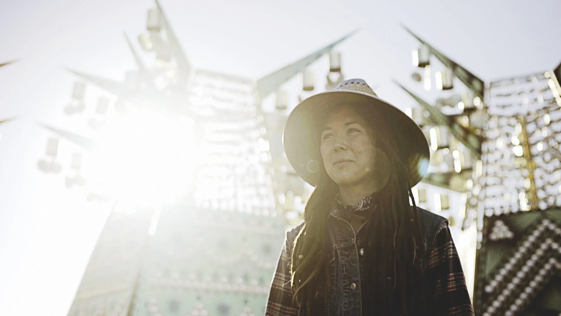 A woman with dreadlocks wearing a wide-brimmed hat and denim jacket stands outdoors with a large Ferris wheel in the background, illuminated by sunlight.