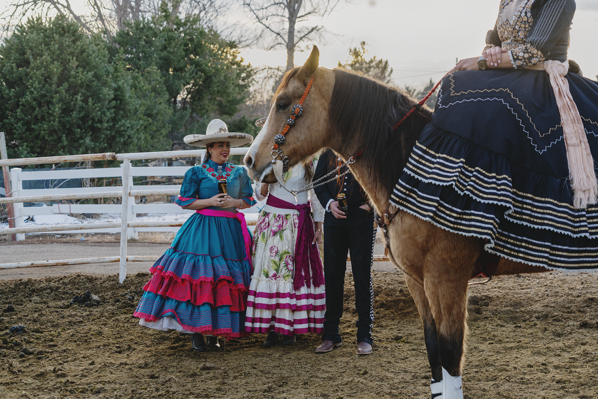 People dressed in traditional Mexican attire gathered around a horse in an outdoor arena drinking Guinness beer.
