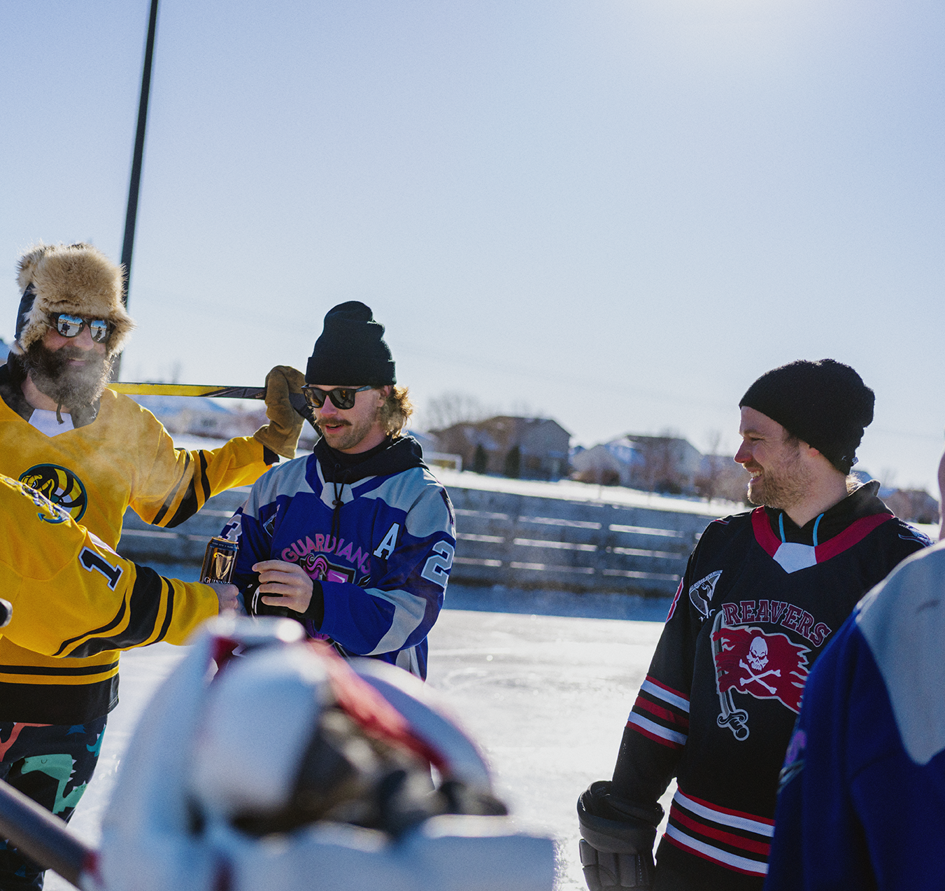 Four men in hockey jerseys and winter gear standing on an outdoor ice rink, smiling and talking on a sunny winter day.