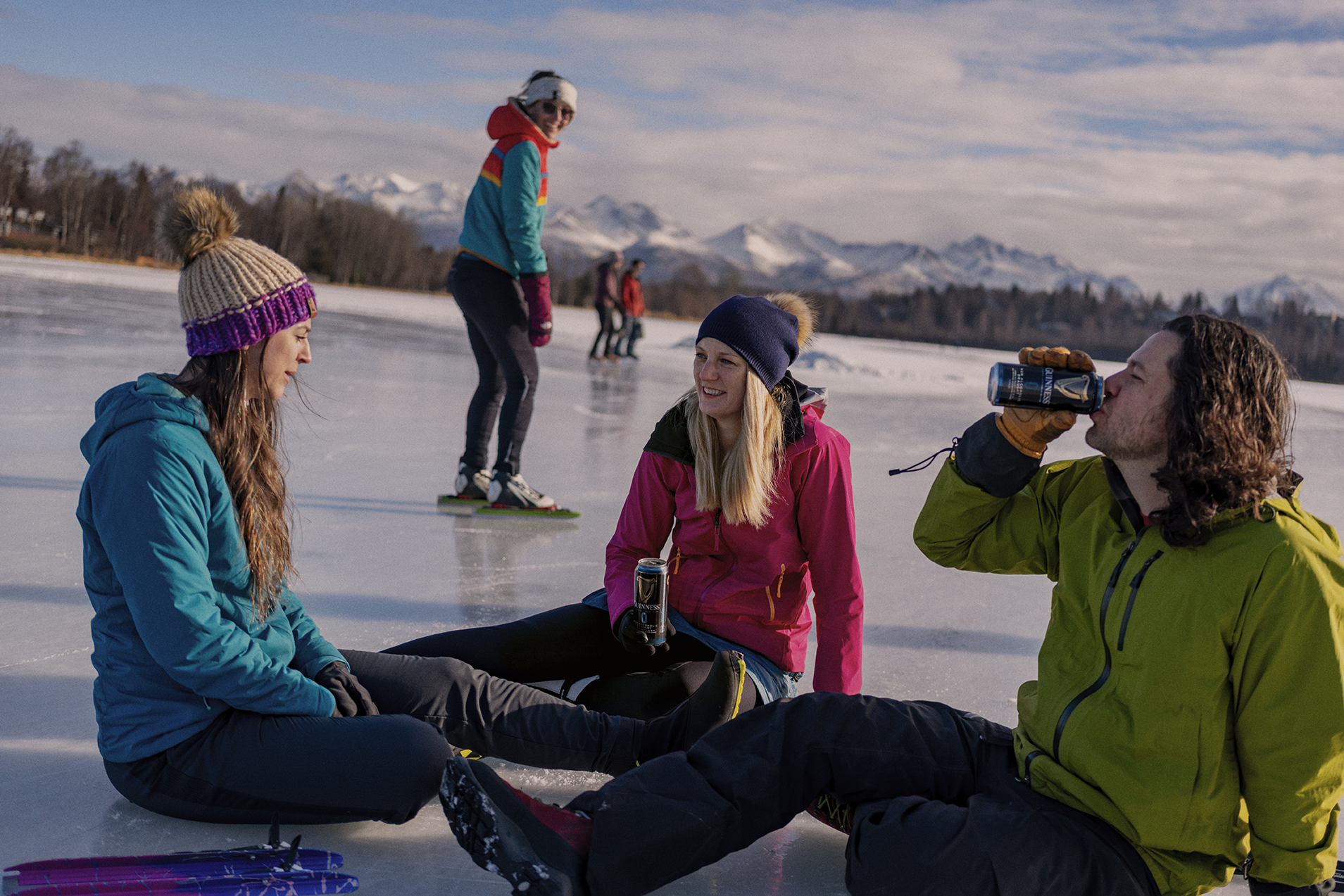 Group of friends relaxing on an ice-covered lake in winter, with snow-capped mountains in the background. Some are sitting on the ice while others are standing or ice skating, drinking Guiness beer, beer, and enjoying the outdoors.