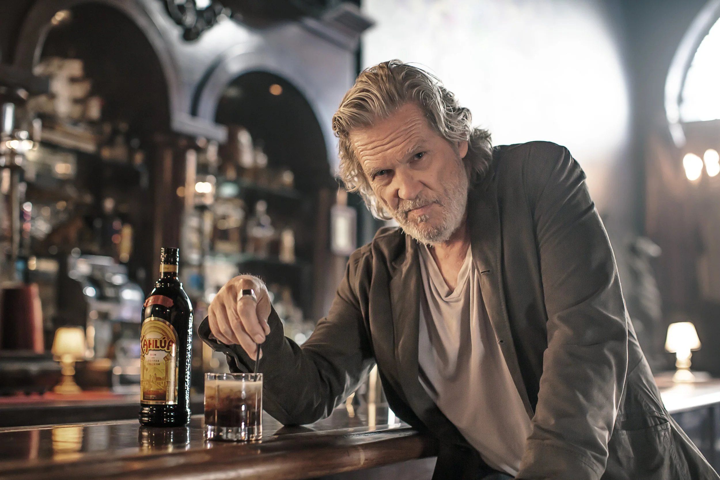 Jeff Bridges is stirring a drink at a bar. A bottle of Ahlúa coffee liqueur and a glass of dark liquid are on the bar counter in front of him. The background features dark wood shelves with various bottles and warm lighting.