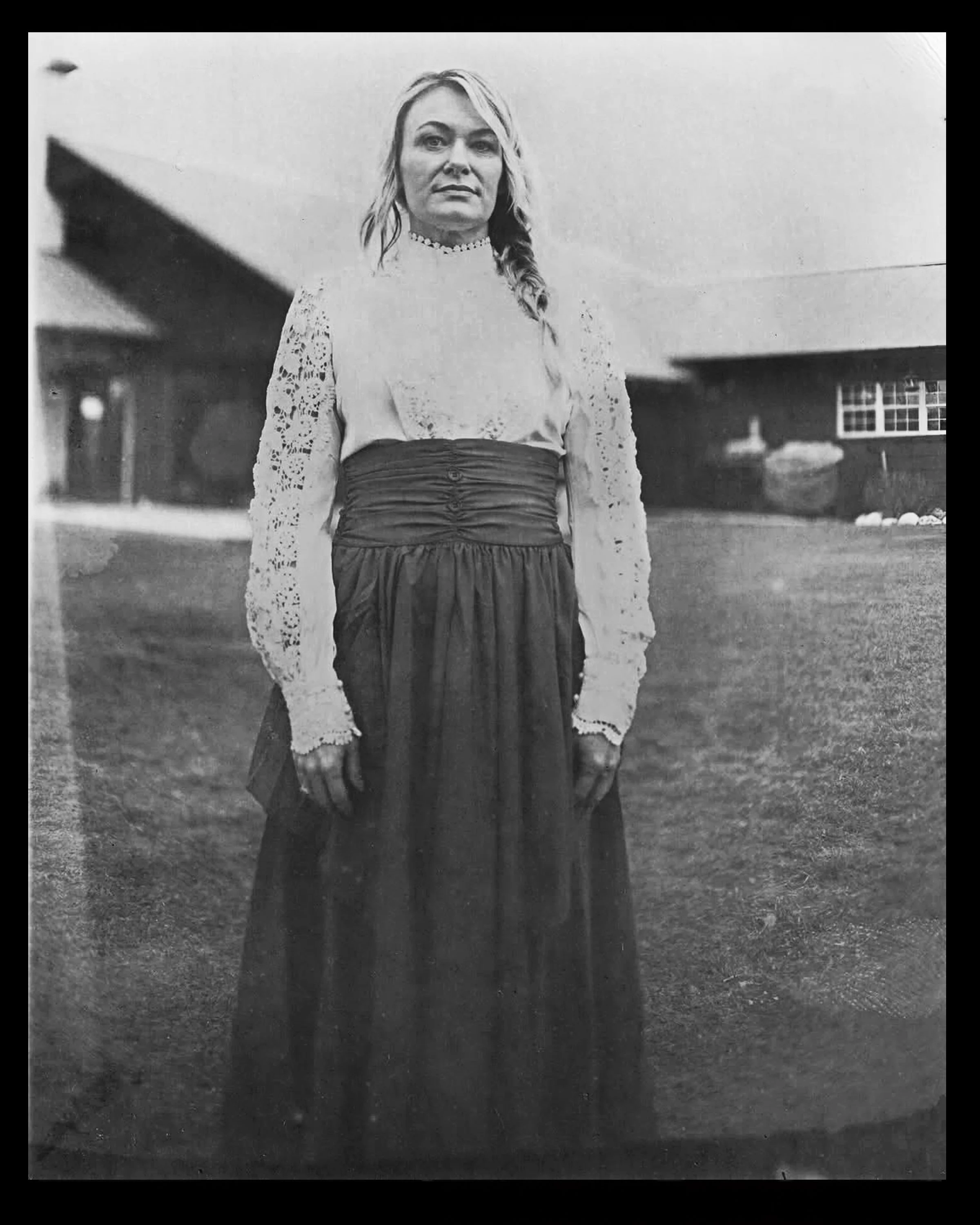 A woman in historical clothing standing outdoors in front of a house with a sloped roof. She has long hair, a calm expression, and is wearing a lace blouse with long sleeves and a high collar, along with a gathered, high-waisted skirt.