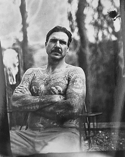 A shirtless man with extensive tattoos on his chest, arms, and shoulders sits outdoors with arms crossed, in a wooded area with trees in the background.