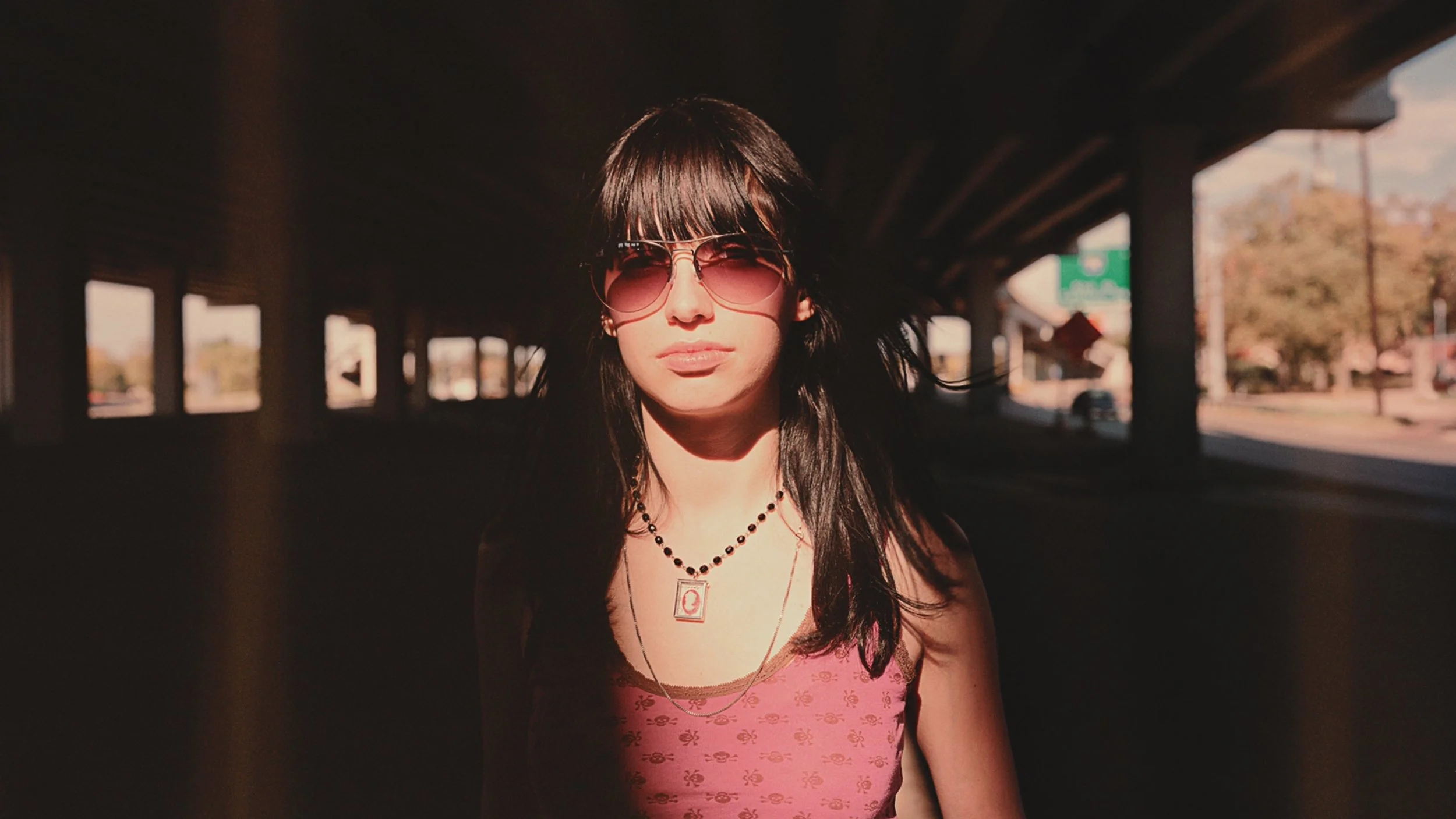 A young woman with dark hair wearing sunglasses, a pink tank top, and layered necklaces stands under an overpass with sunlight casting shadows on her face.