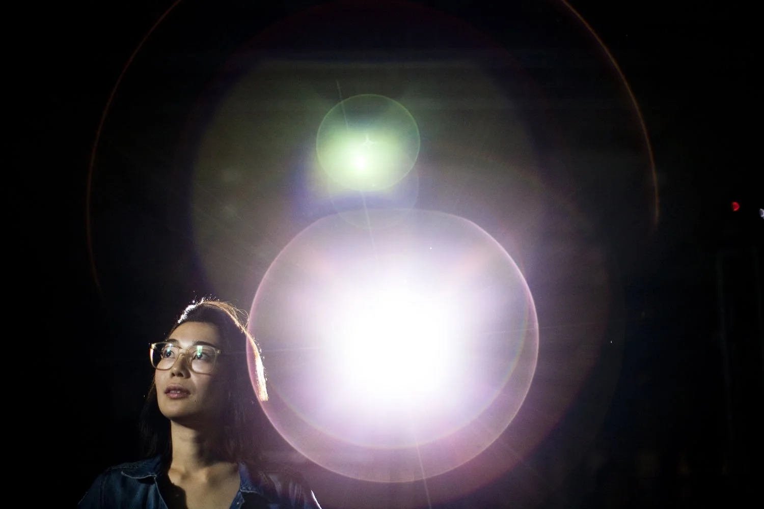 A young woman with glasses and dark hair looking upward against a dark background with bright lens flare effects.
