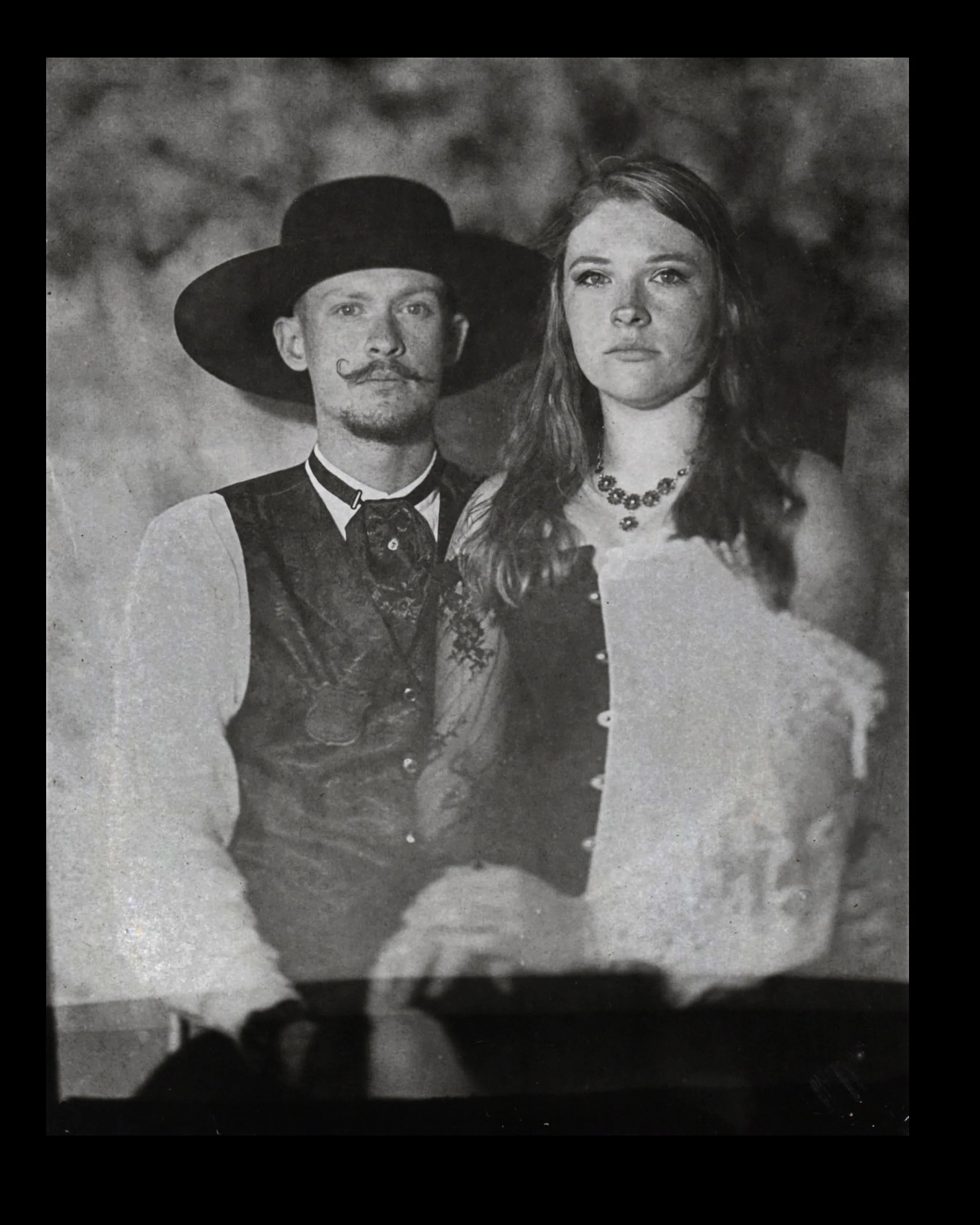 A black-and-white photo of a man with a large hat, styled mustache and beard, standing behind a woman with long wavy hair, wearing a necklace. Both are dressed in vintage-style clothing.