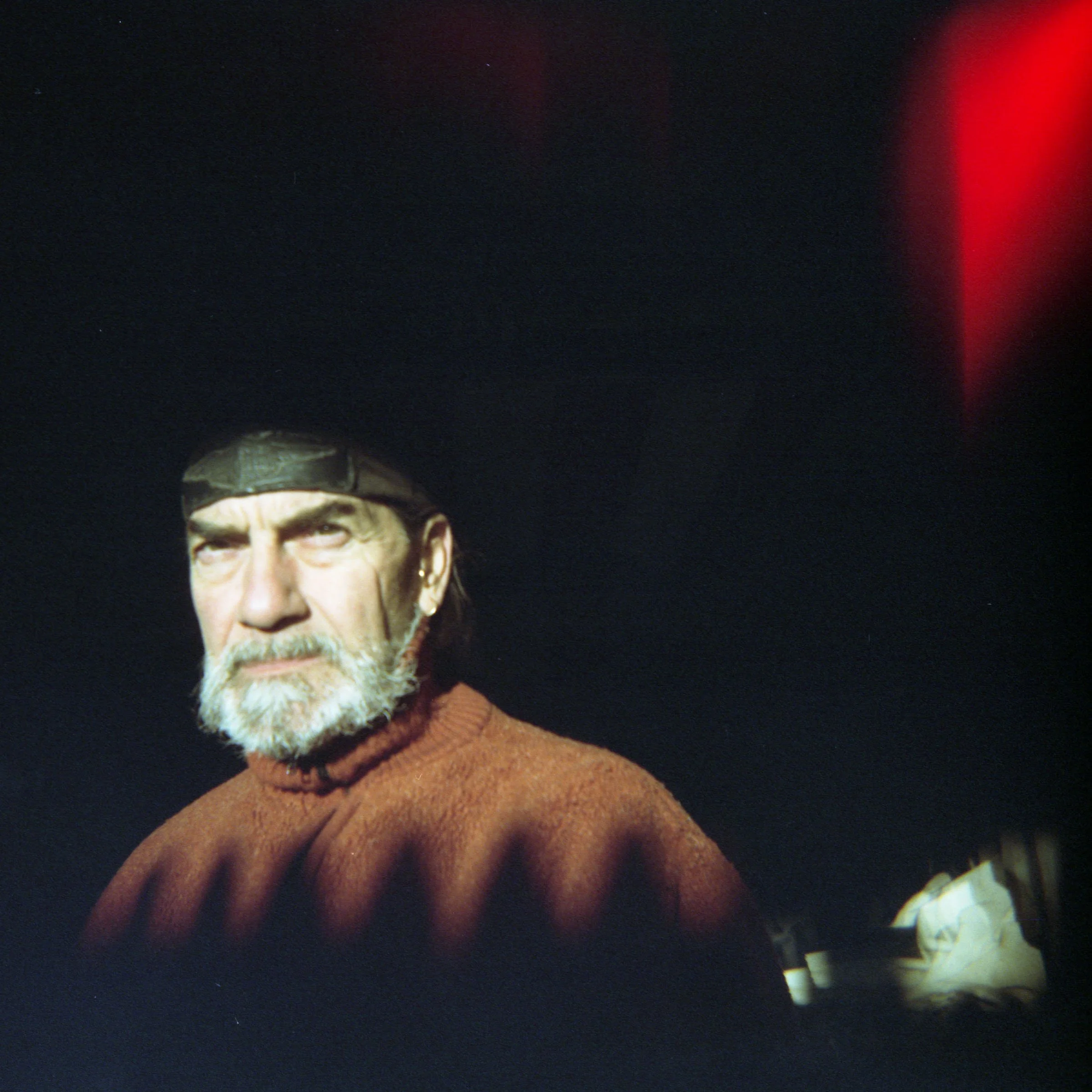 A man with gray hair, beard, wearing a black cap and a rust-colored sweater against a dark background with red lighting.