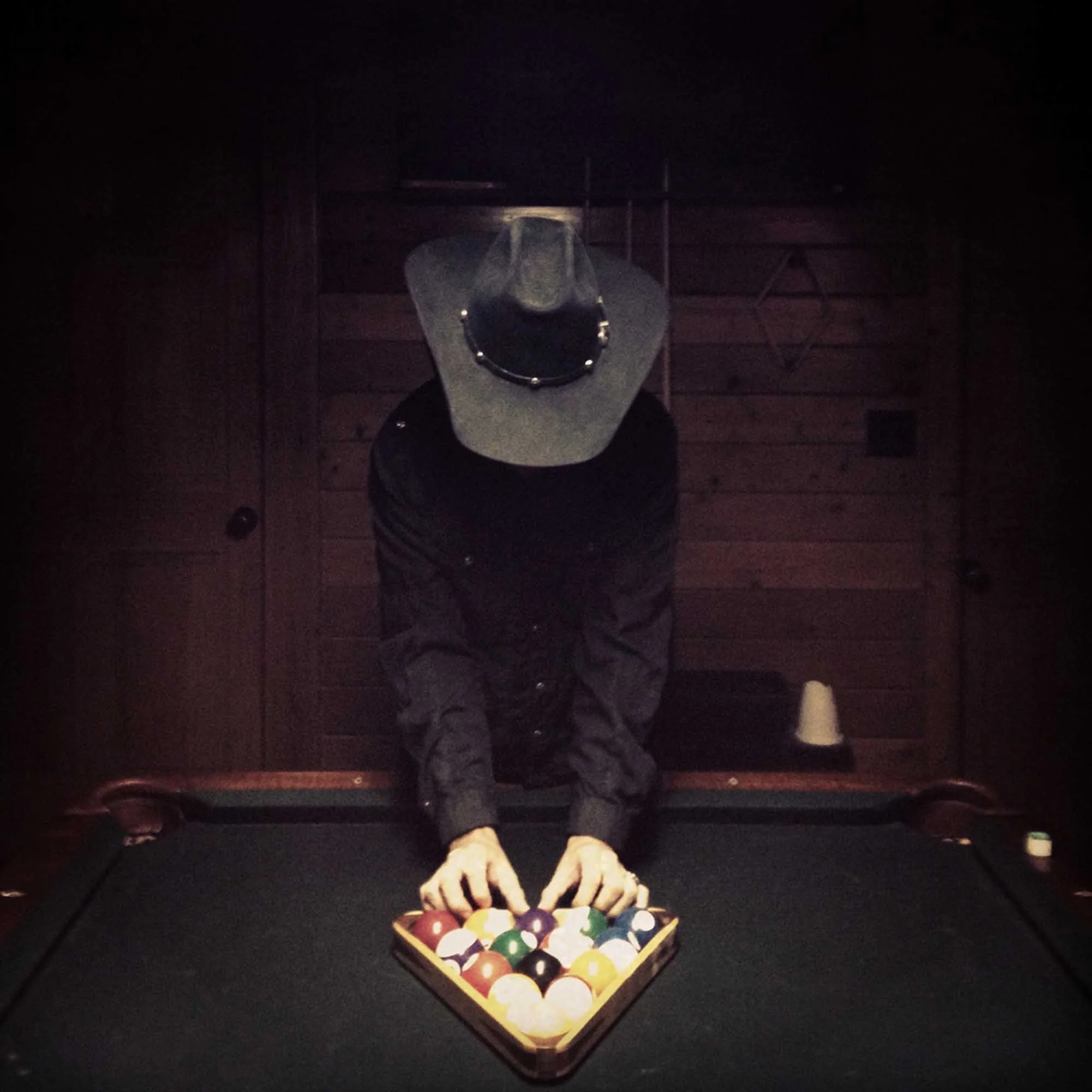A person wearing a cowboy hat and black jacket, leaning over a pool table, preparing to take a shot, with a wooden wall in the background.