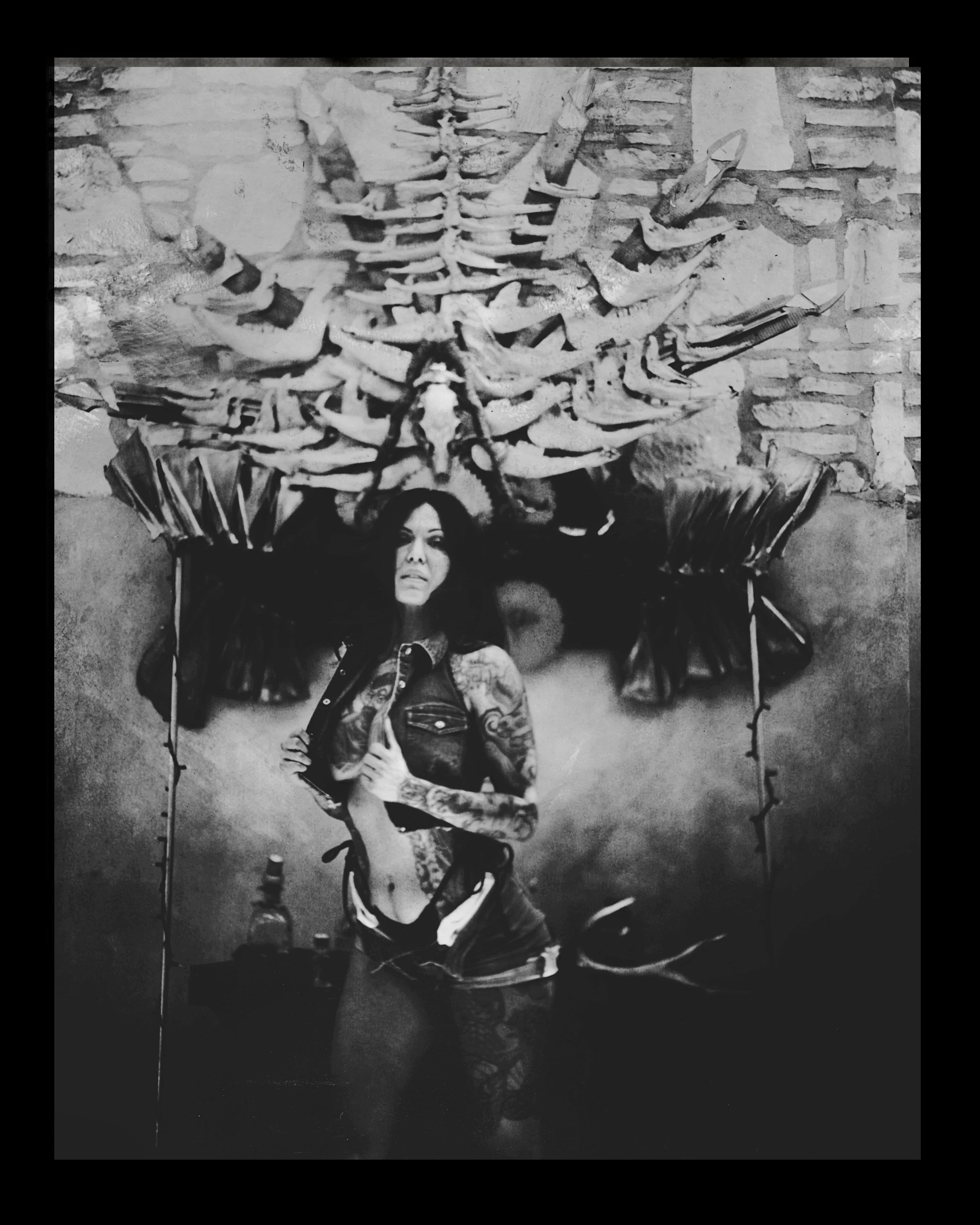 A woman with tattoos poses in front of a wall with a large skeleton mounted above her, and large butterfly wings on either side of her. The photo is black and white.