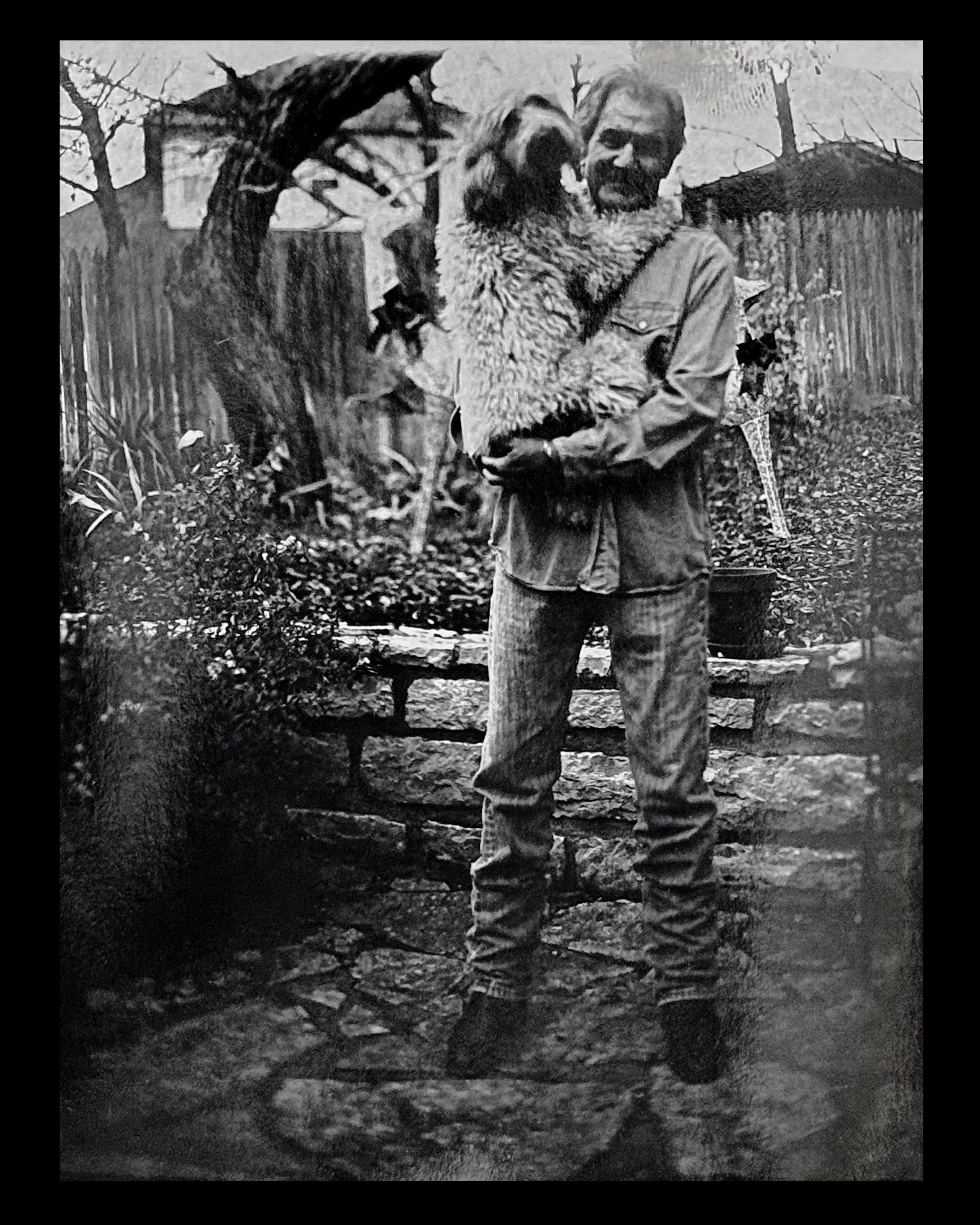 A black and white photo of a man holding a dog in a backyard garden with a stone pathway, wooden fence, trees, and plants.