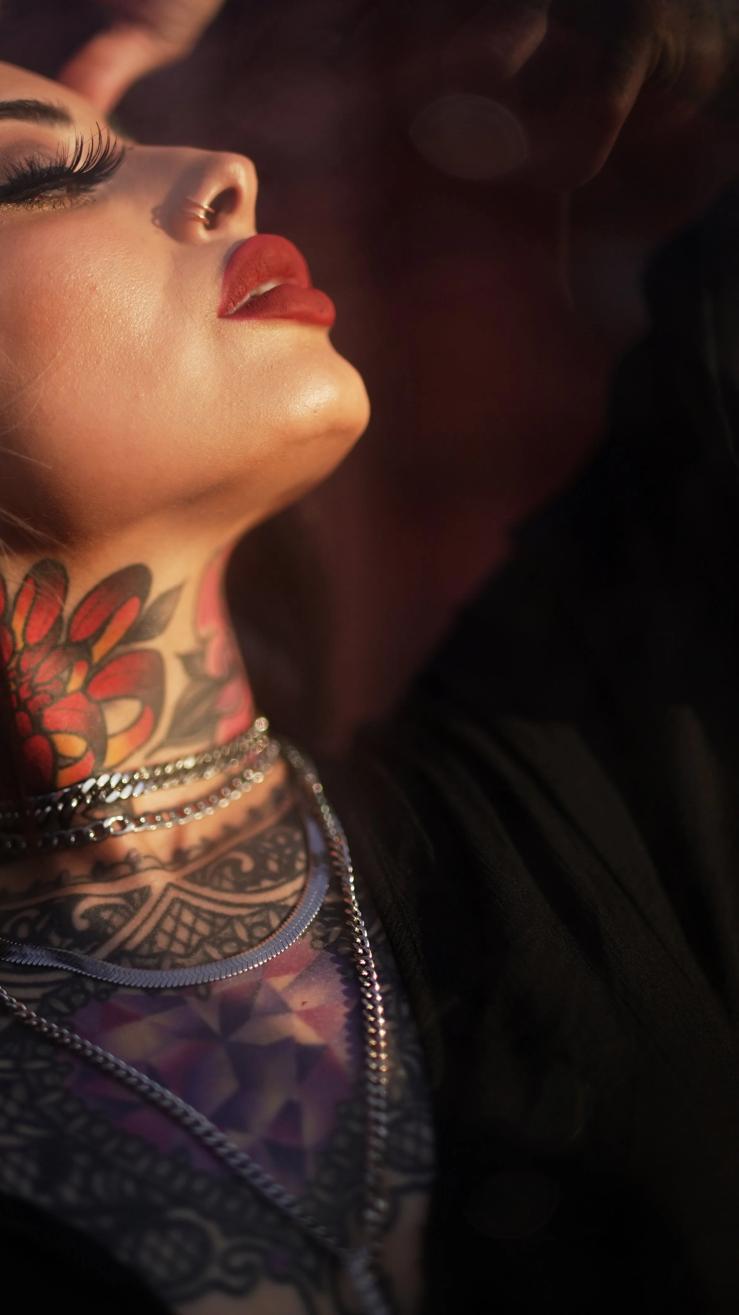 Close-up of a woman with colorful tattoos on her neck, wearing lipstick, and adorned with layered necklaces.