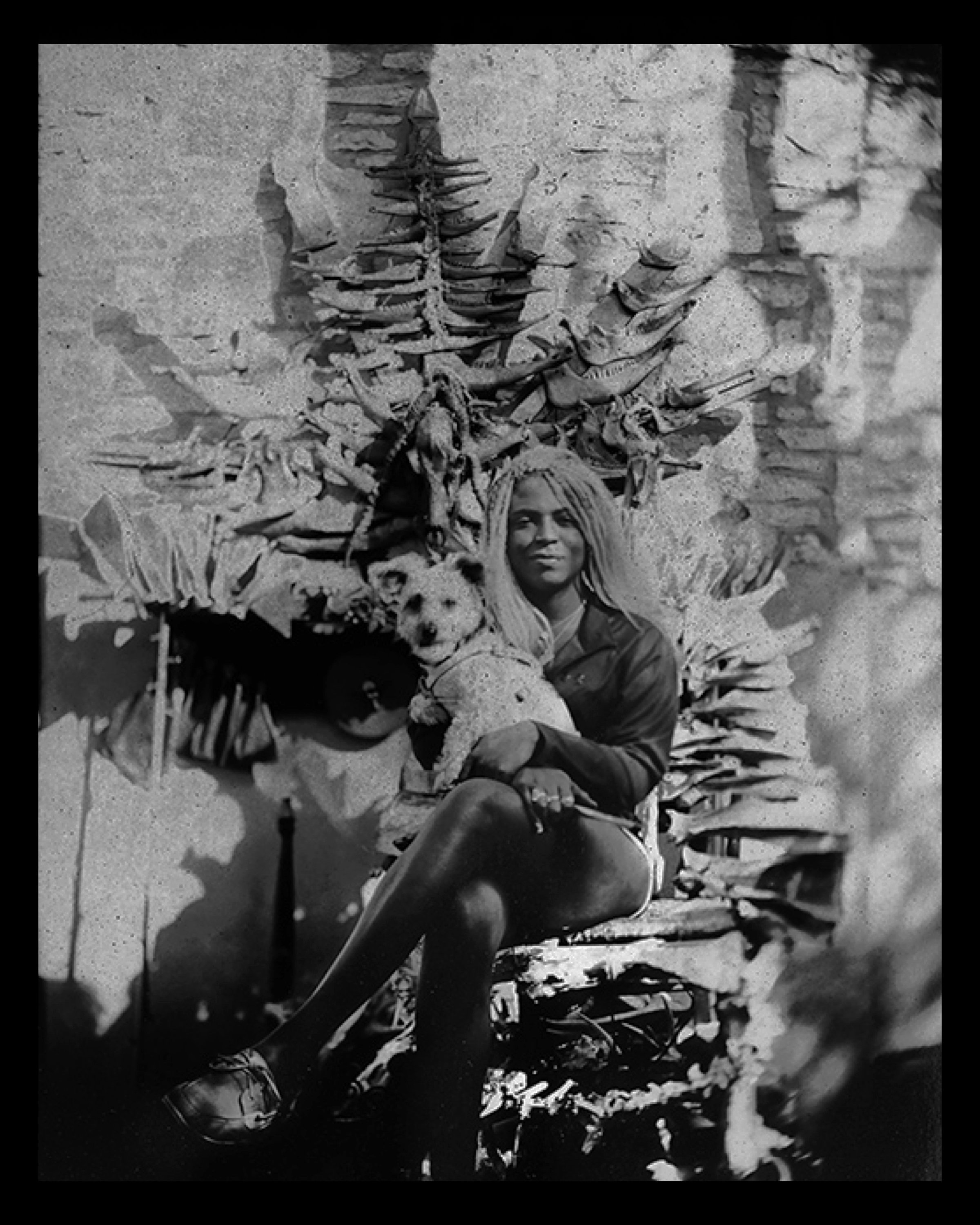 A person with dreadlocks sitting in front of a wall decorated with animal bones and skulls, holding a teddy bear.