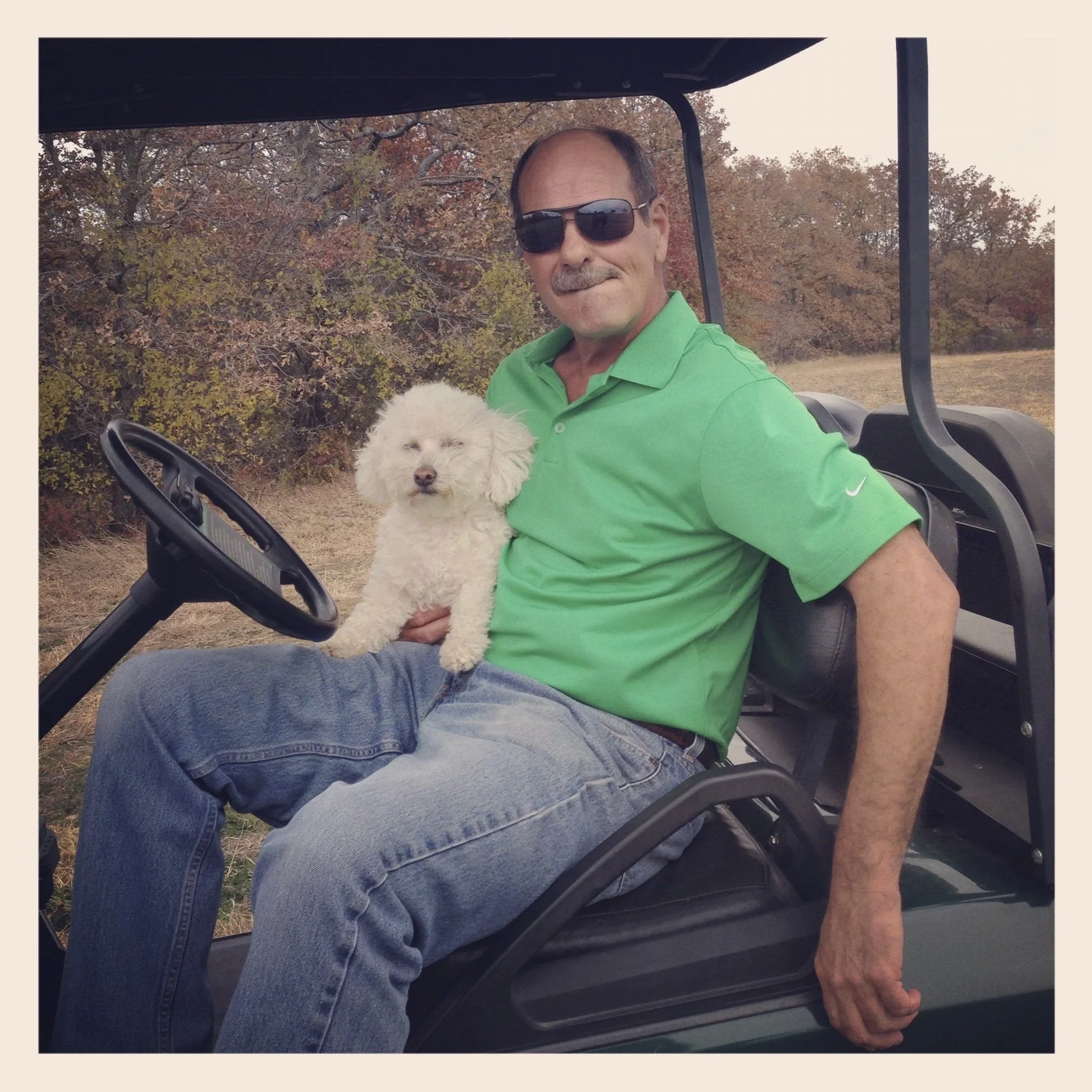 A man sitting on a golf cart holding a small white dog outside in a rural setting with trees and grass in the background.