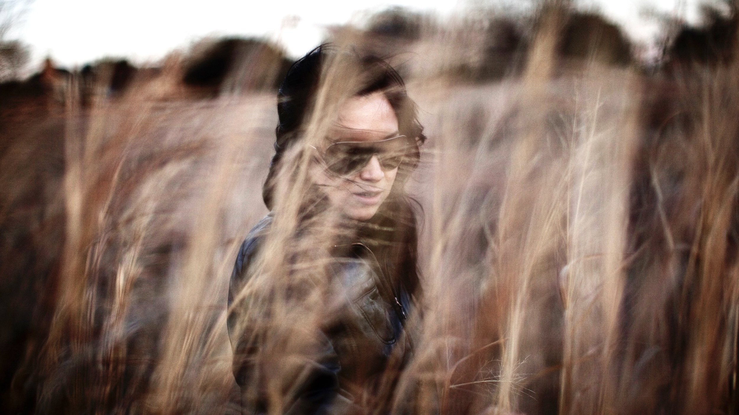 A woman with dark hair wearing sunglasses and a leather jacket standing among tall dried grasses.