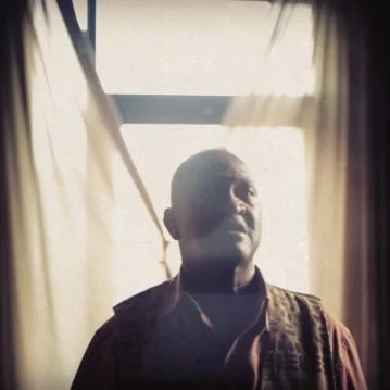 A man stands behind semi-transparent curtains, with sunlight coming through a window behind him.