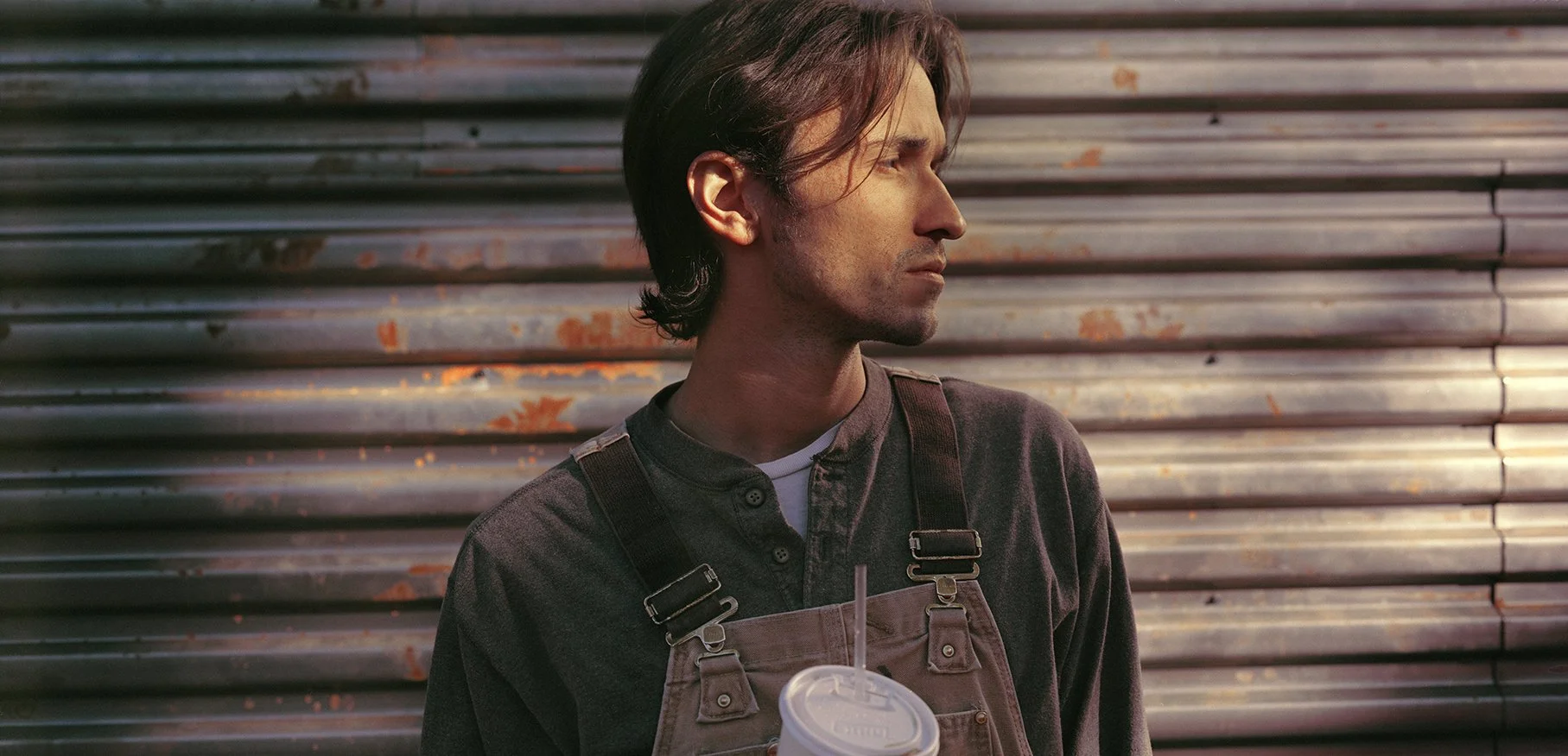 Side profile of a young man with dark hair, wearing a dark shirt and brown overalls, holding a cup with a straw, standing in front of a metal, rusted corrugated wall.