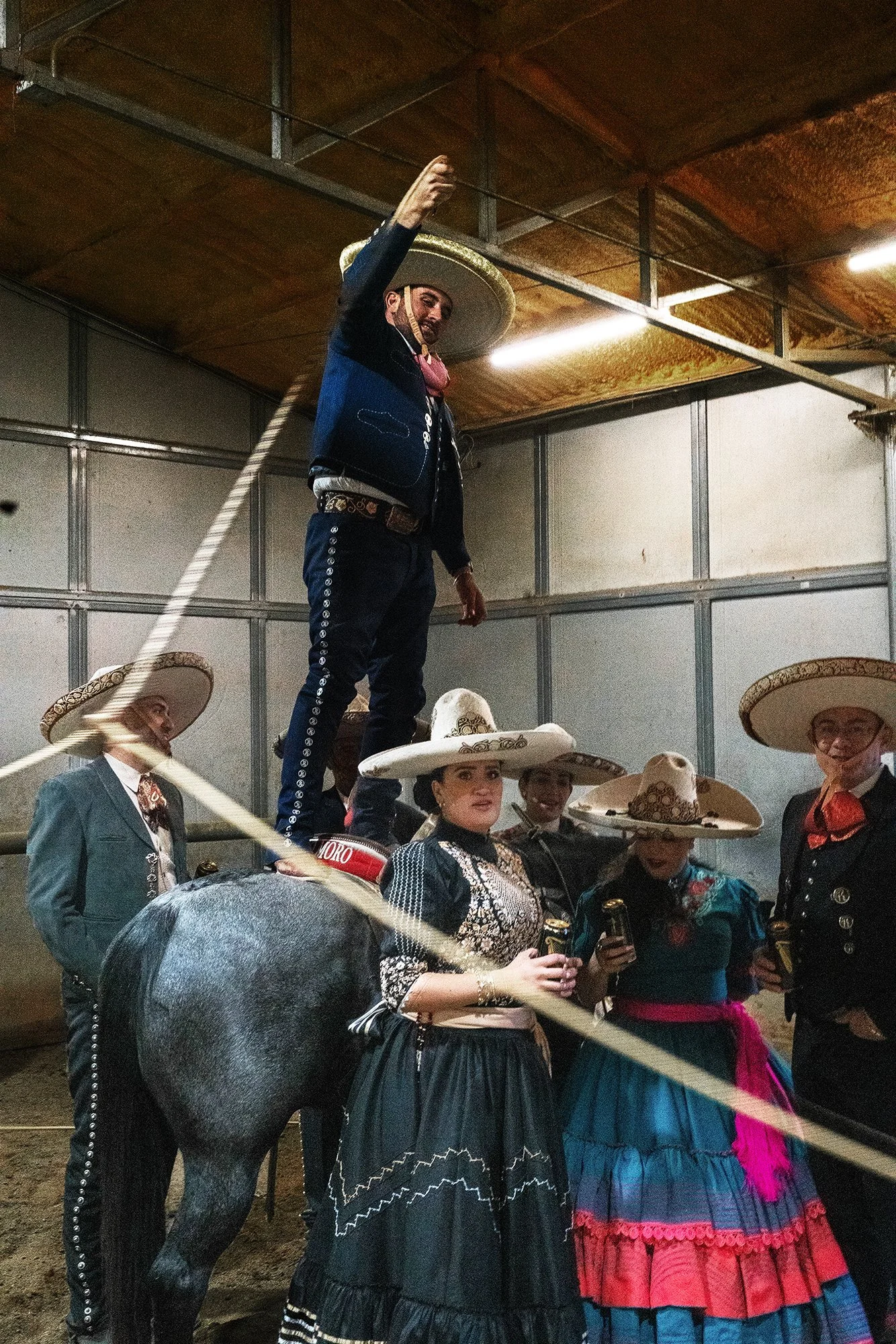 A man dressed as a matador standing on a horse, surrounded by women in traditional Mexican dresses and sombreros, inside a rustic building drinking Guiness beer.