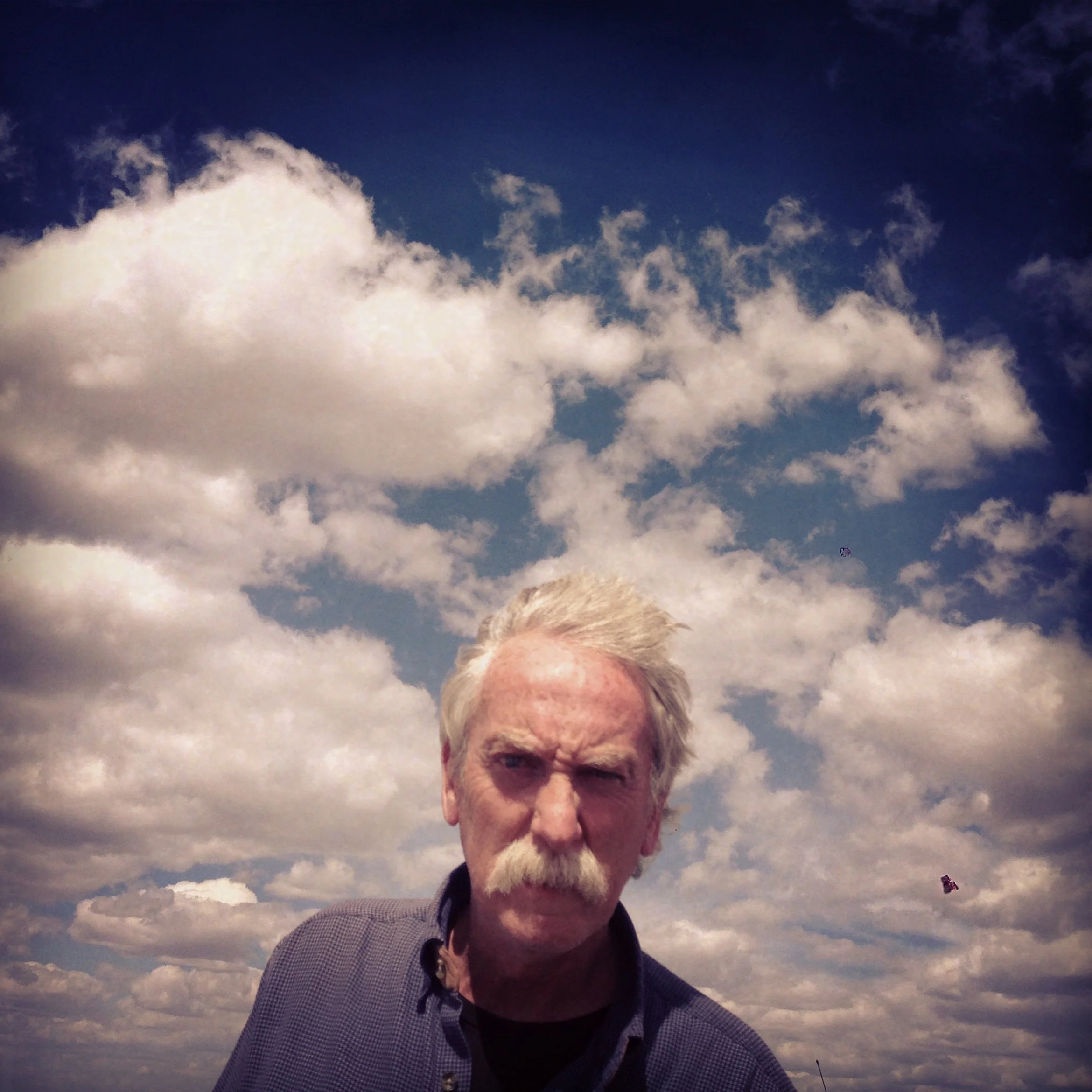 A man with white hair and a mustache is outdoors under a partly cloudy sky, looking directly at the camera.