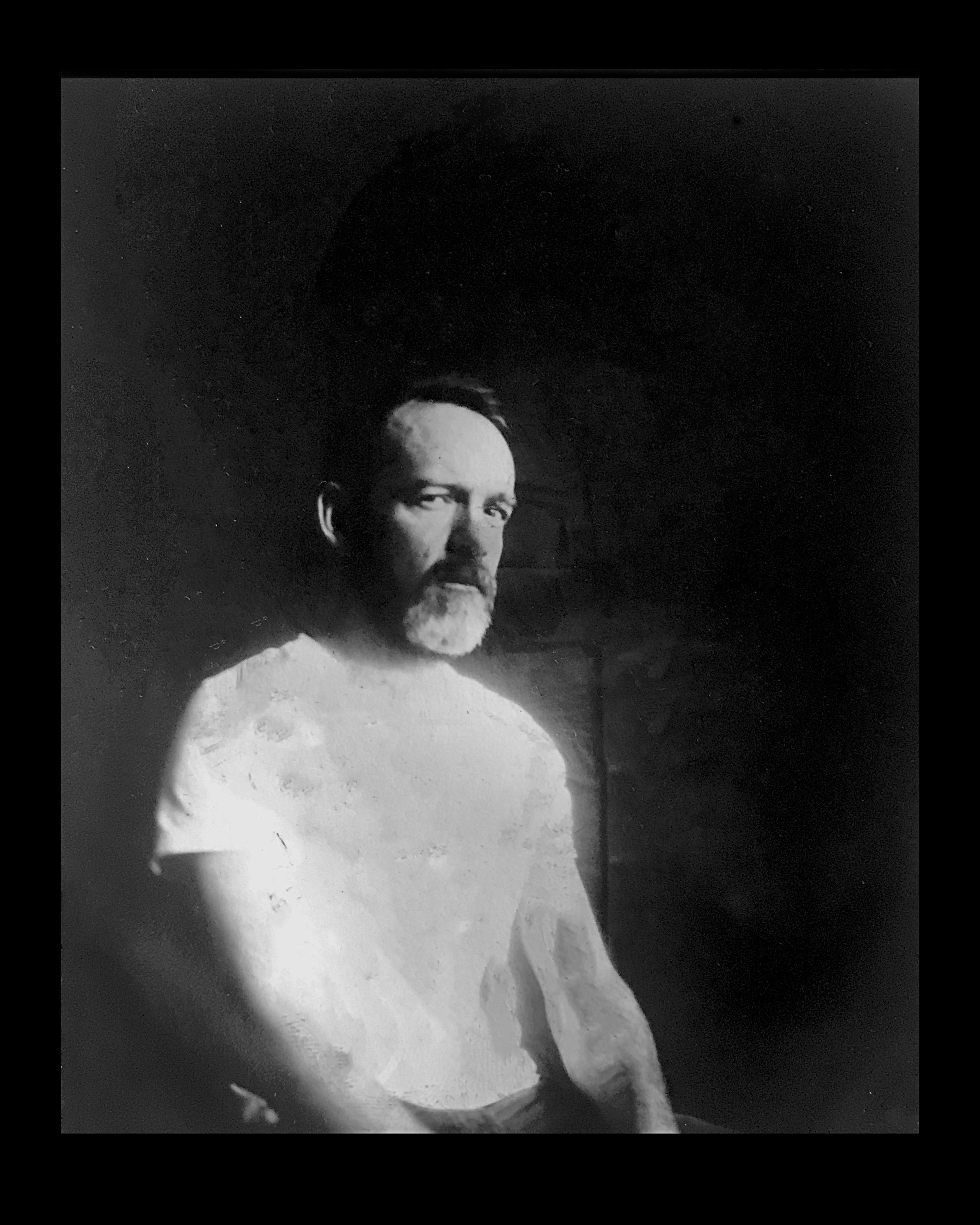 Black and white photograph of a man with a beard, sitting and looking at the camera, in a dimly lit room.