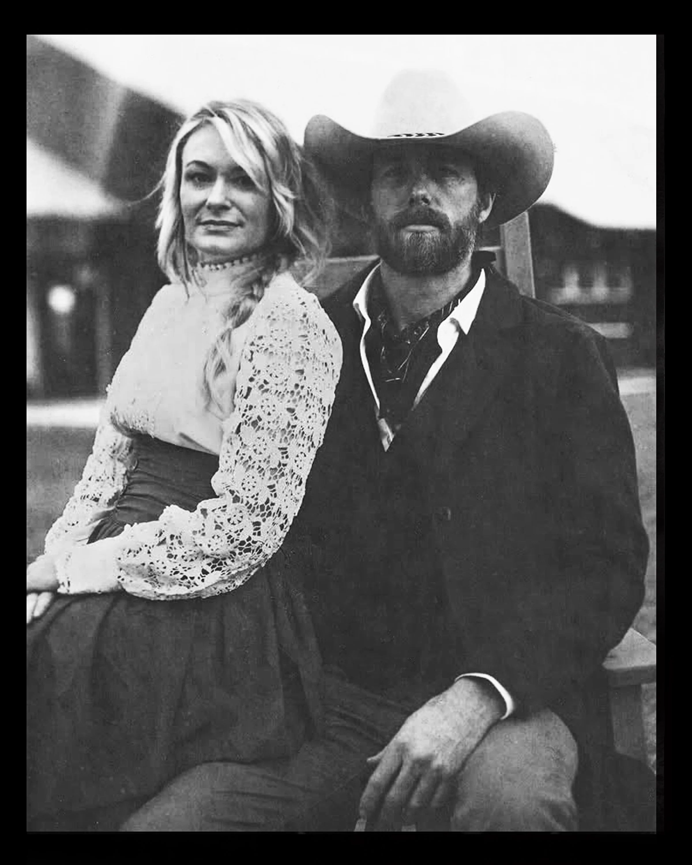 Black and white photo of a man wearing a cowboy hat and a woman with long wavy hair in a lace blouse, sitting outdoors in a rural setting.