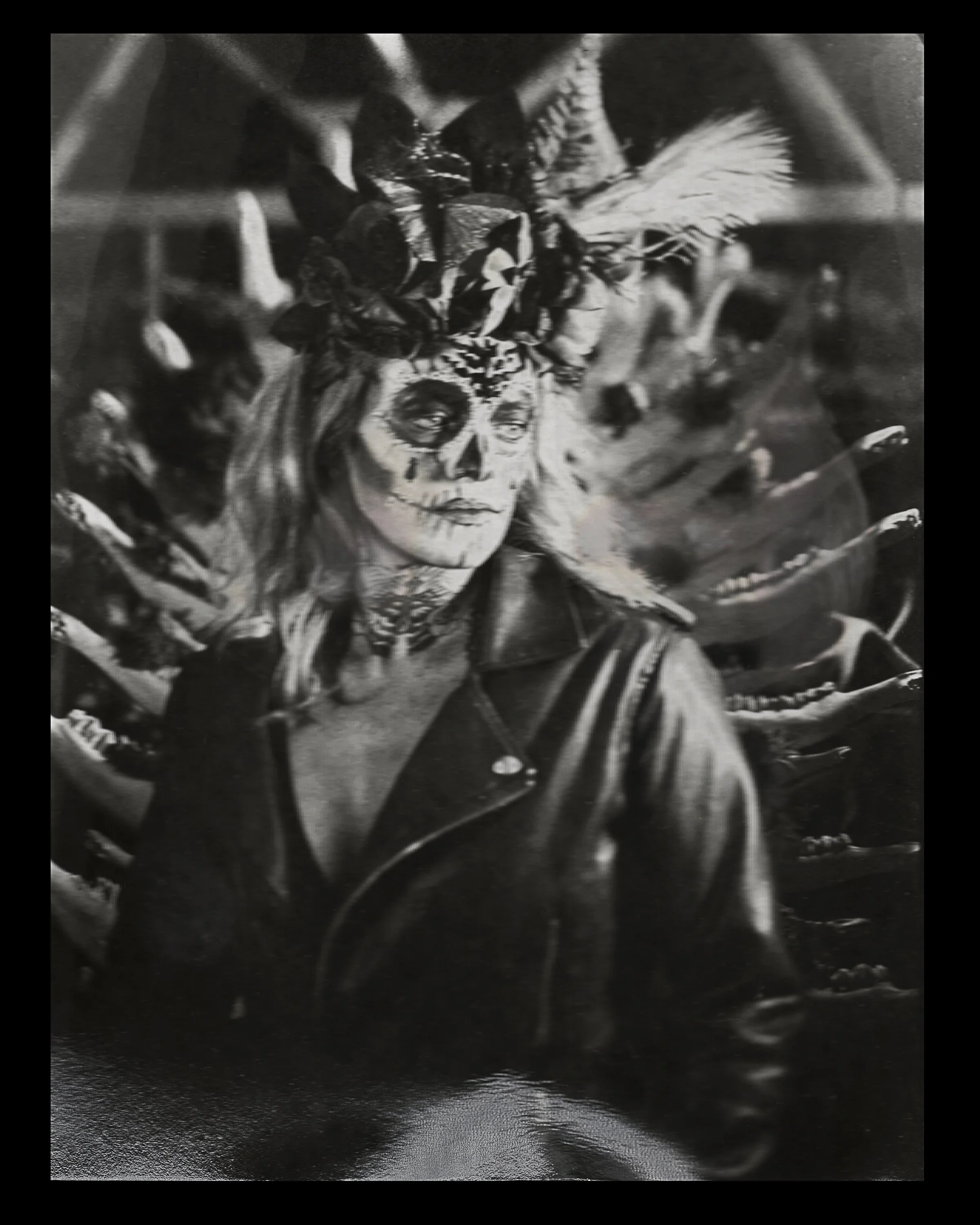 Black and white photograph of a woman with face paint resembling a sugar skull, wearing a headdress with feathers and flowers, and a leather jacket, surrounded by skulls and skeletal imagery.