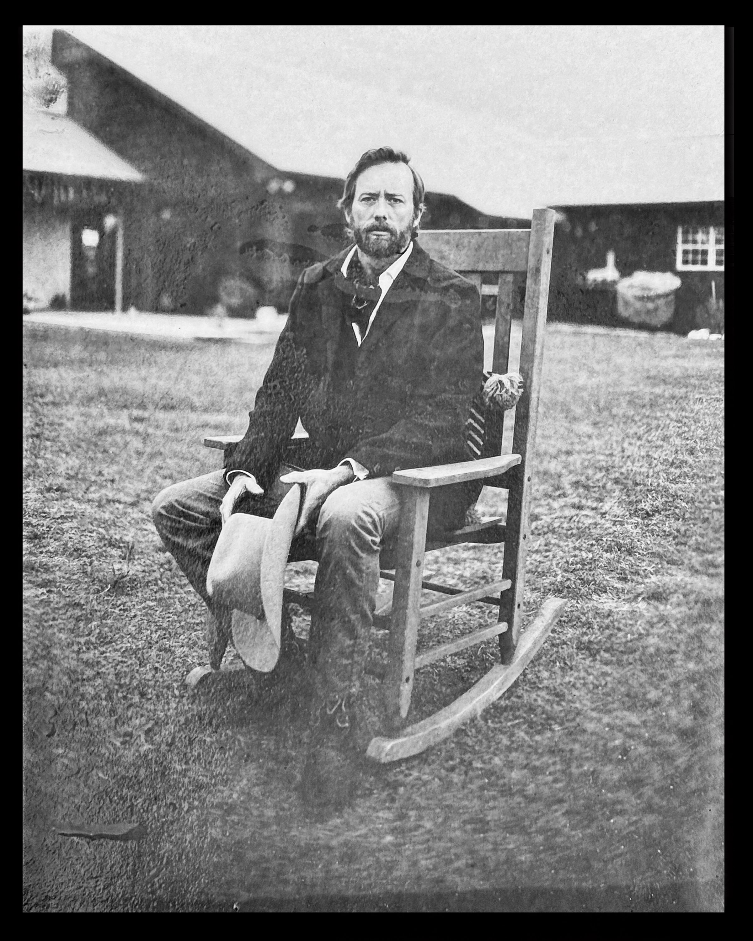 A black and white photograph of a man sitting on a wooden rocking chair outdoors, holding a hat, with a house and a lawn in the background.