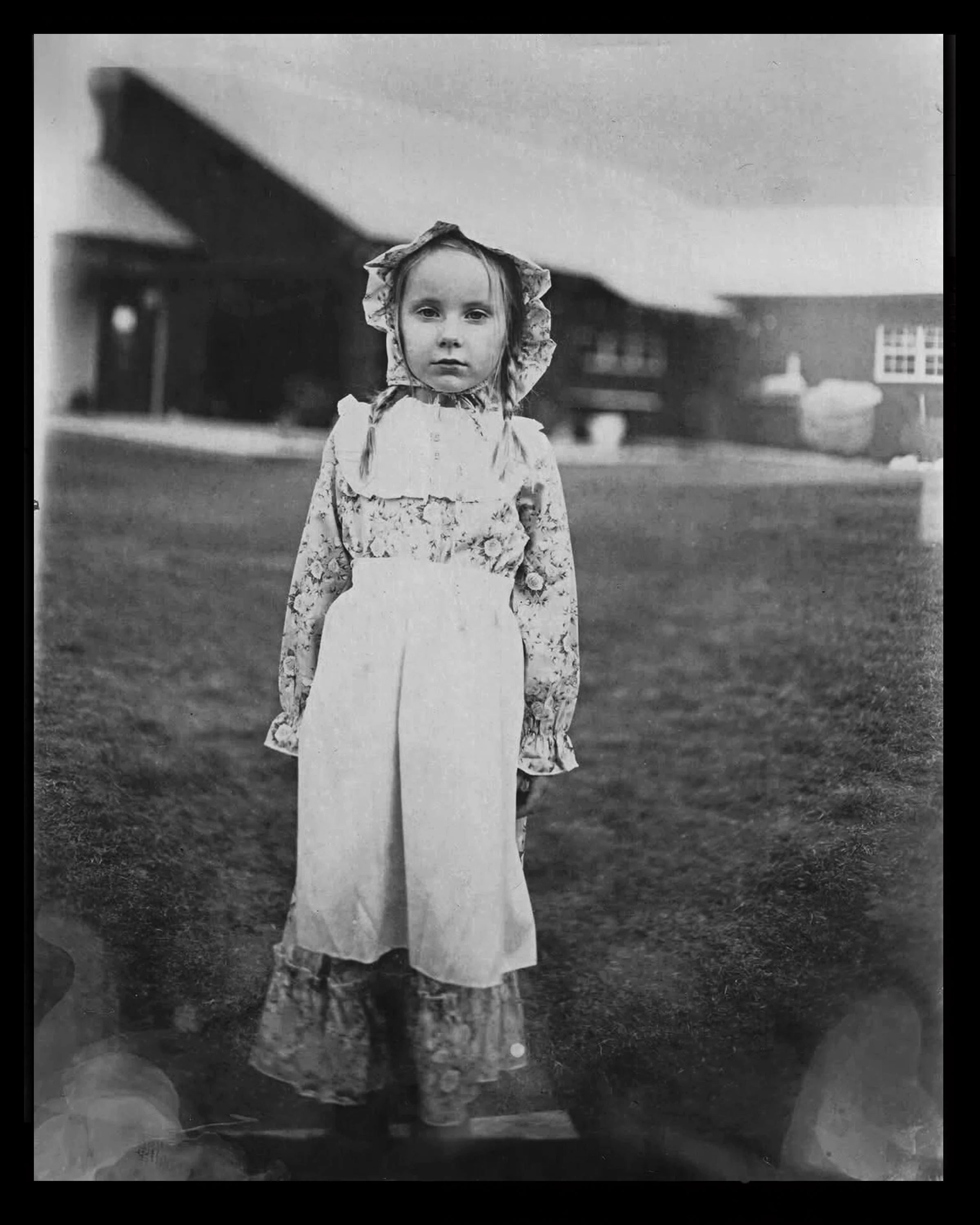 Black and white photograph of a young girl standing outdoors, wearing vintage clothing including a bonnet, floral blouse, high-waisted pants, and patterned shoes, with a large barn or farmhouse in the background.