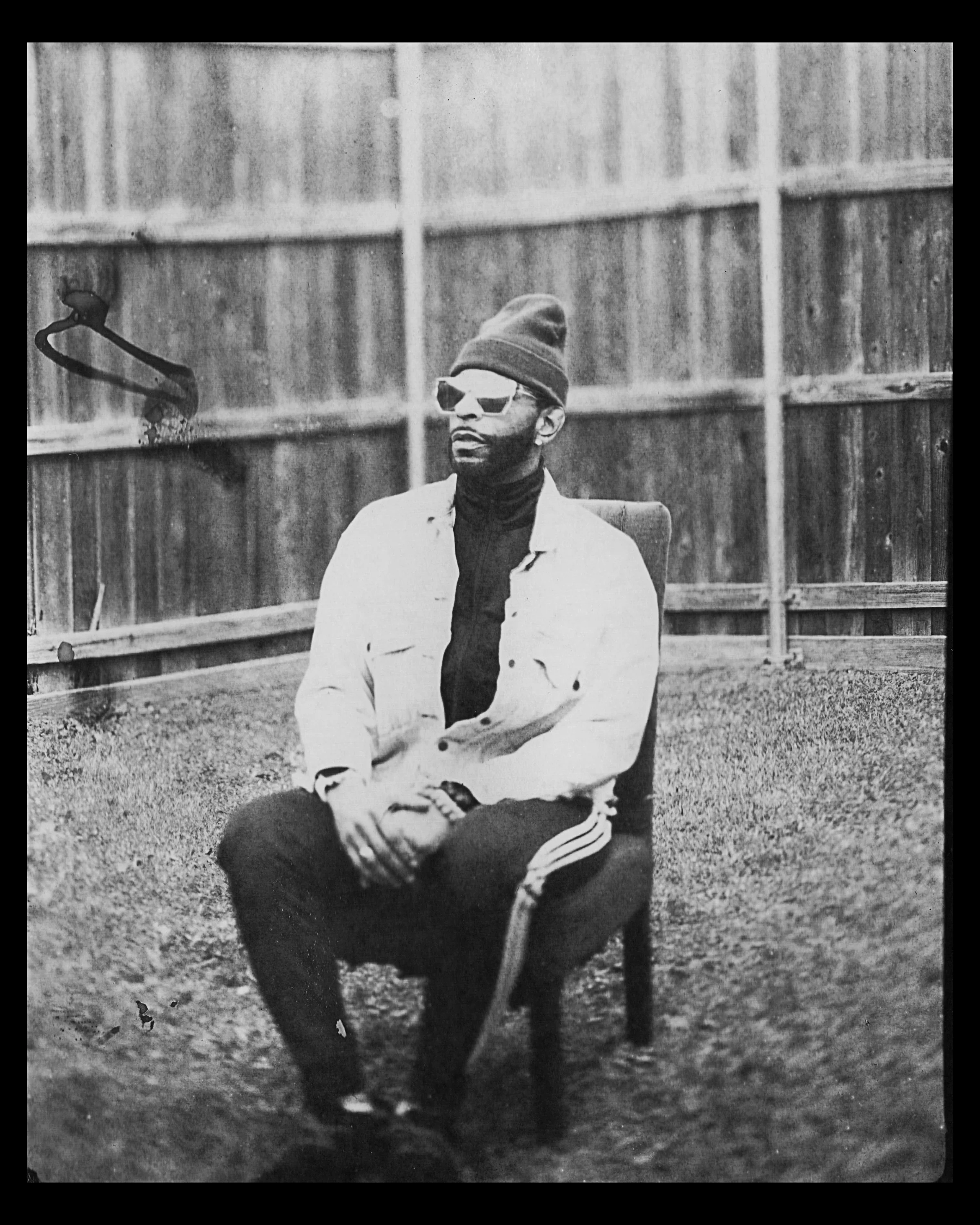 A black and white photo of a man sitting on a chair outdoors, wearing sunglasses, a beanie, a light-colored jacket over a dark turtleneck, and striped track pants, with a wooden fence in the background.