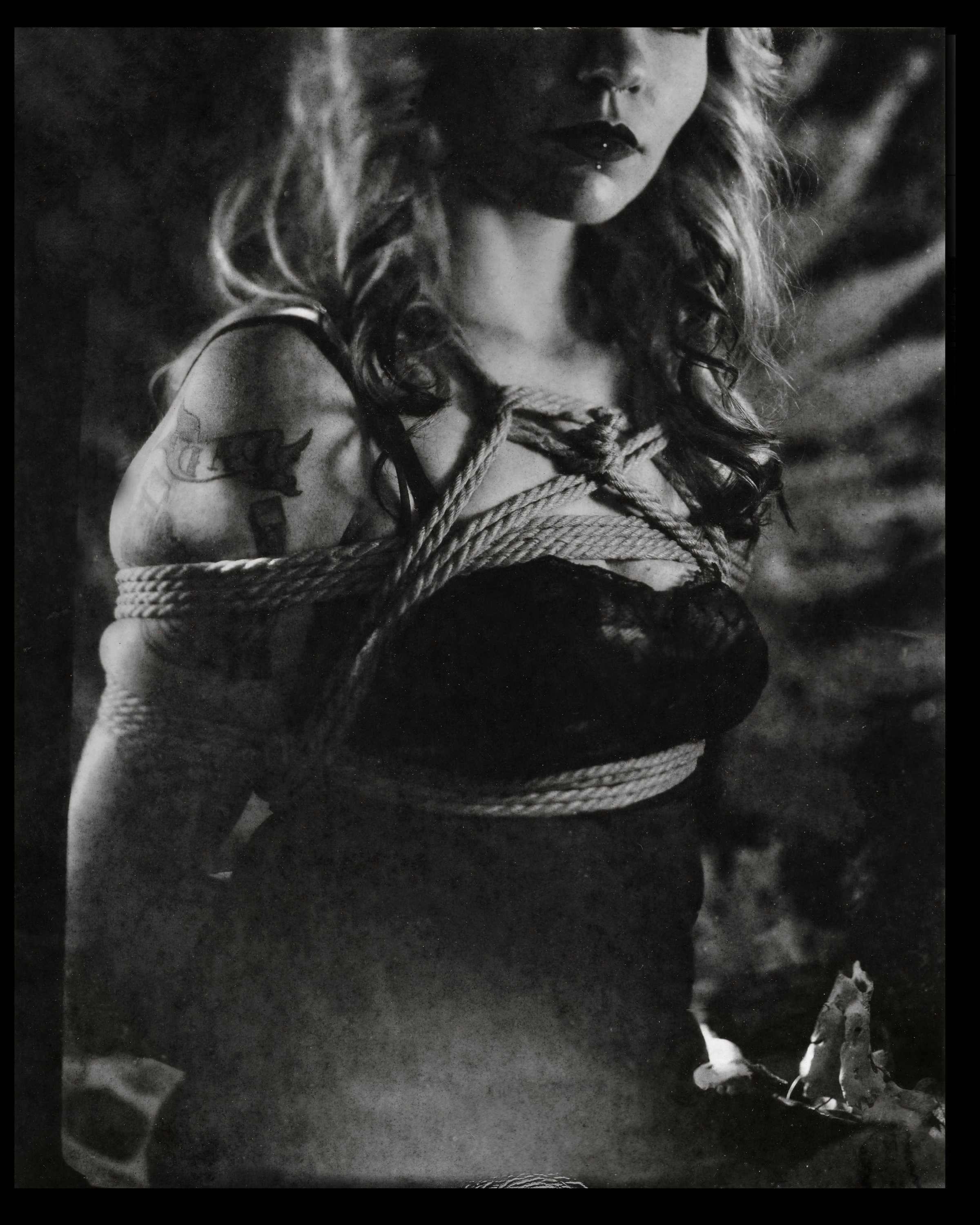 A black and white photograph of a woman with long, wavy hair and dark lipstick, her upper body bound tightly with ropes. She has tattoos on her arm and is wearing a sleeveless top.