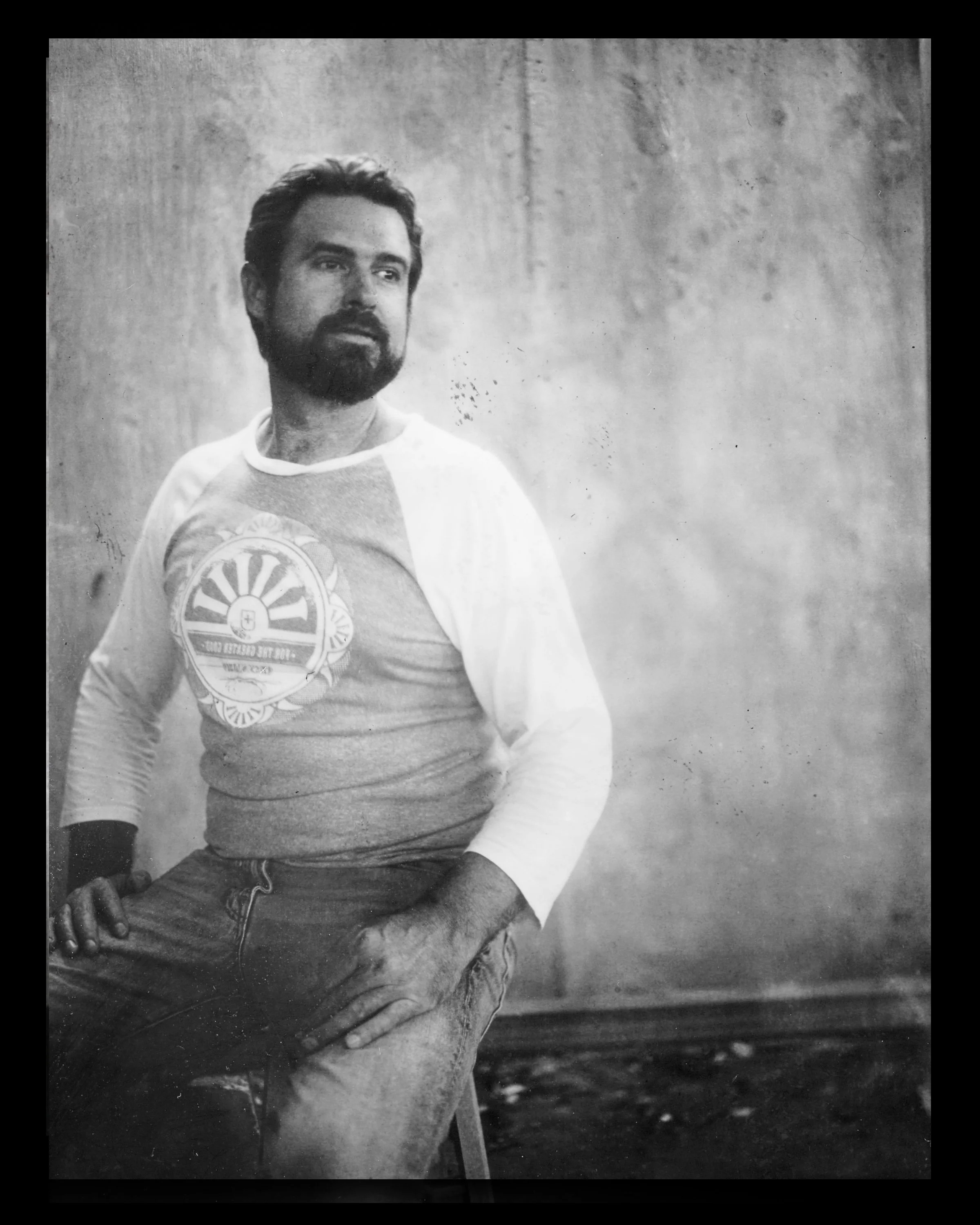 A black and white photo of a man with a beard, sitting with one hand on his knee, wearing a baseball-type shirt with sleeves and jeans, against a plain wall background.