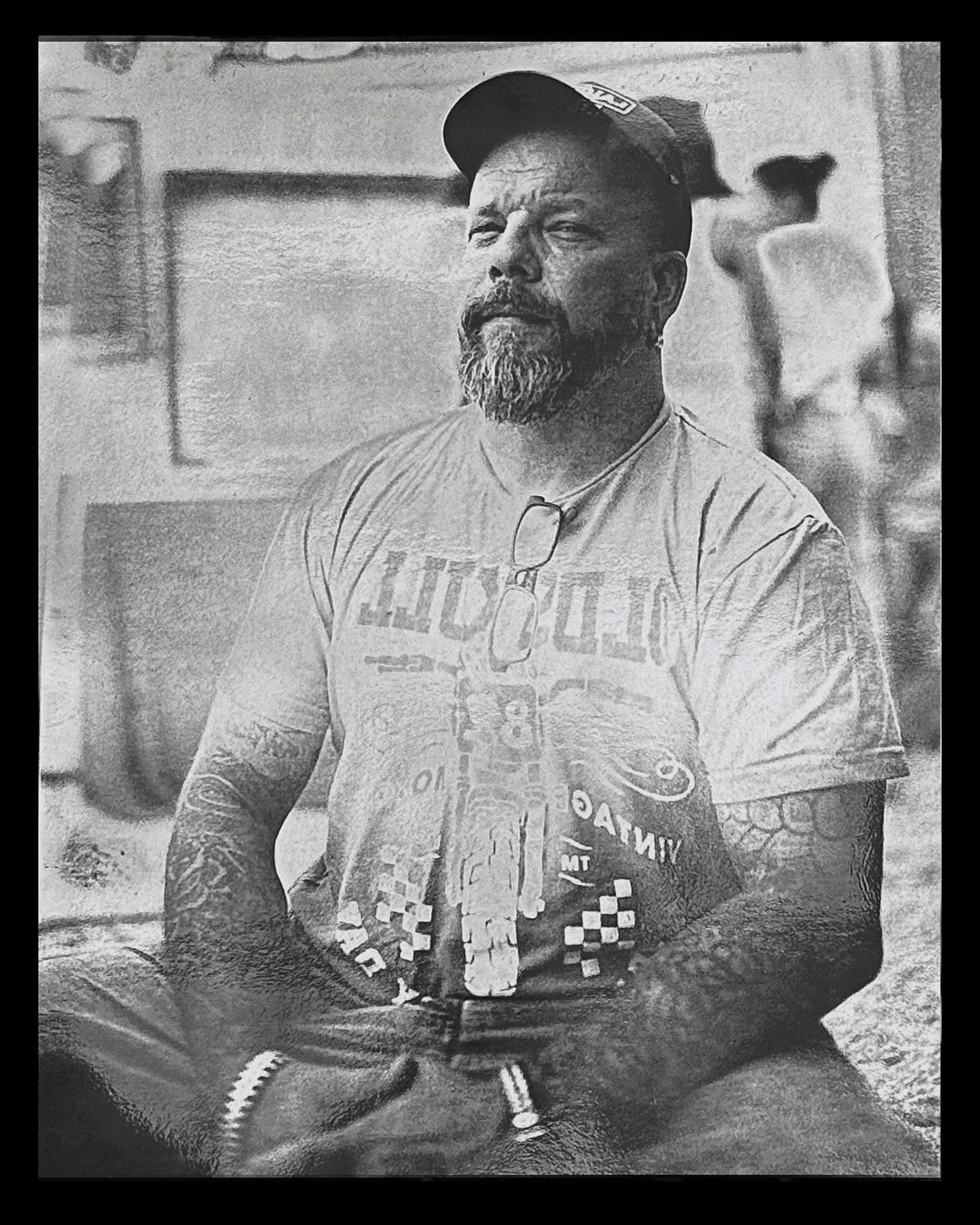 A man with a beard and tattoos, sitting and looking slightly to the side, wearing a baseball cap and a t-shirt with graphic design.