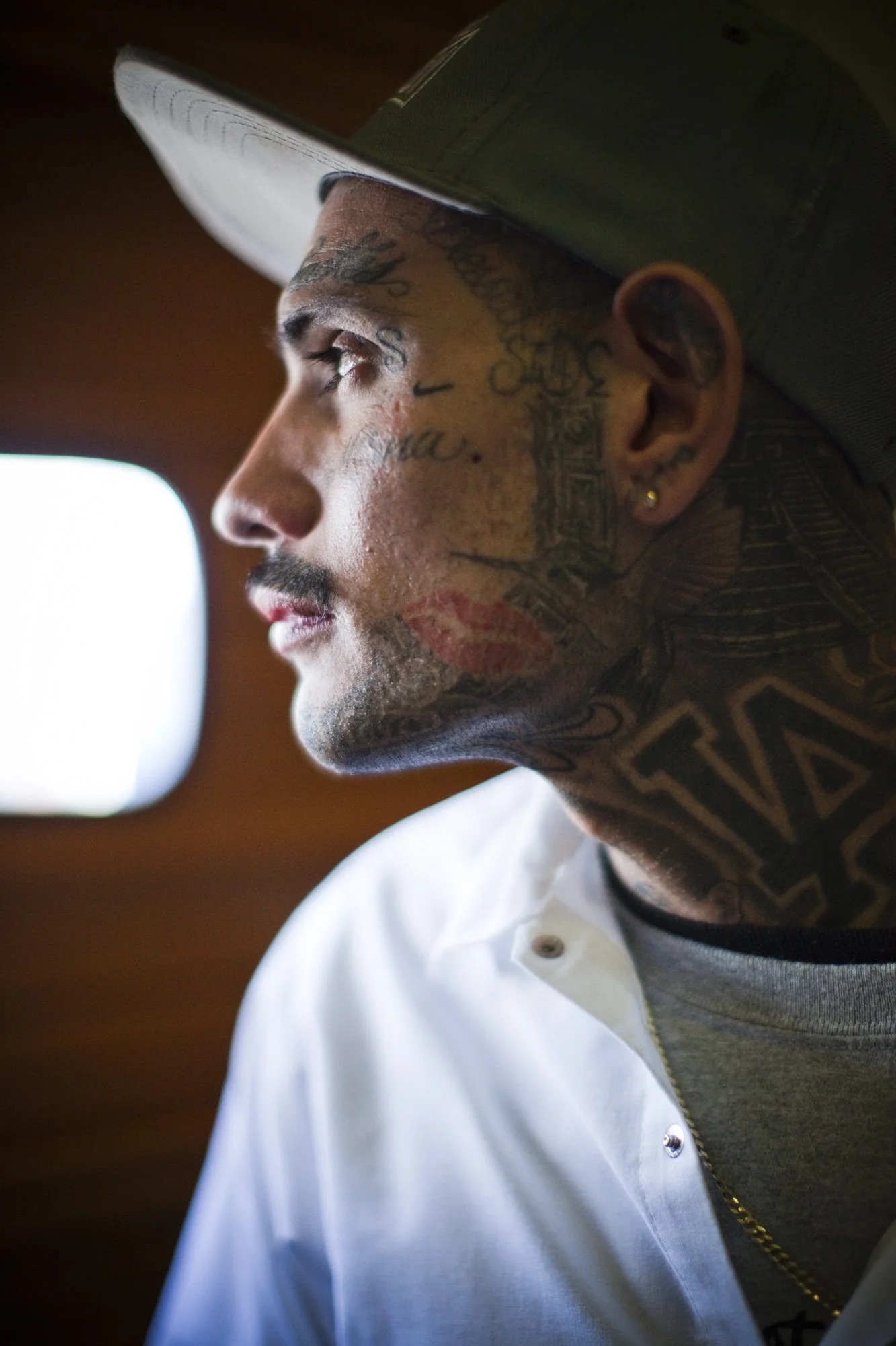 Close-up side profile of a tattooed young man wearing a white shirt and a cap, with a blurred background.