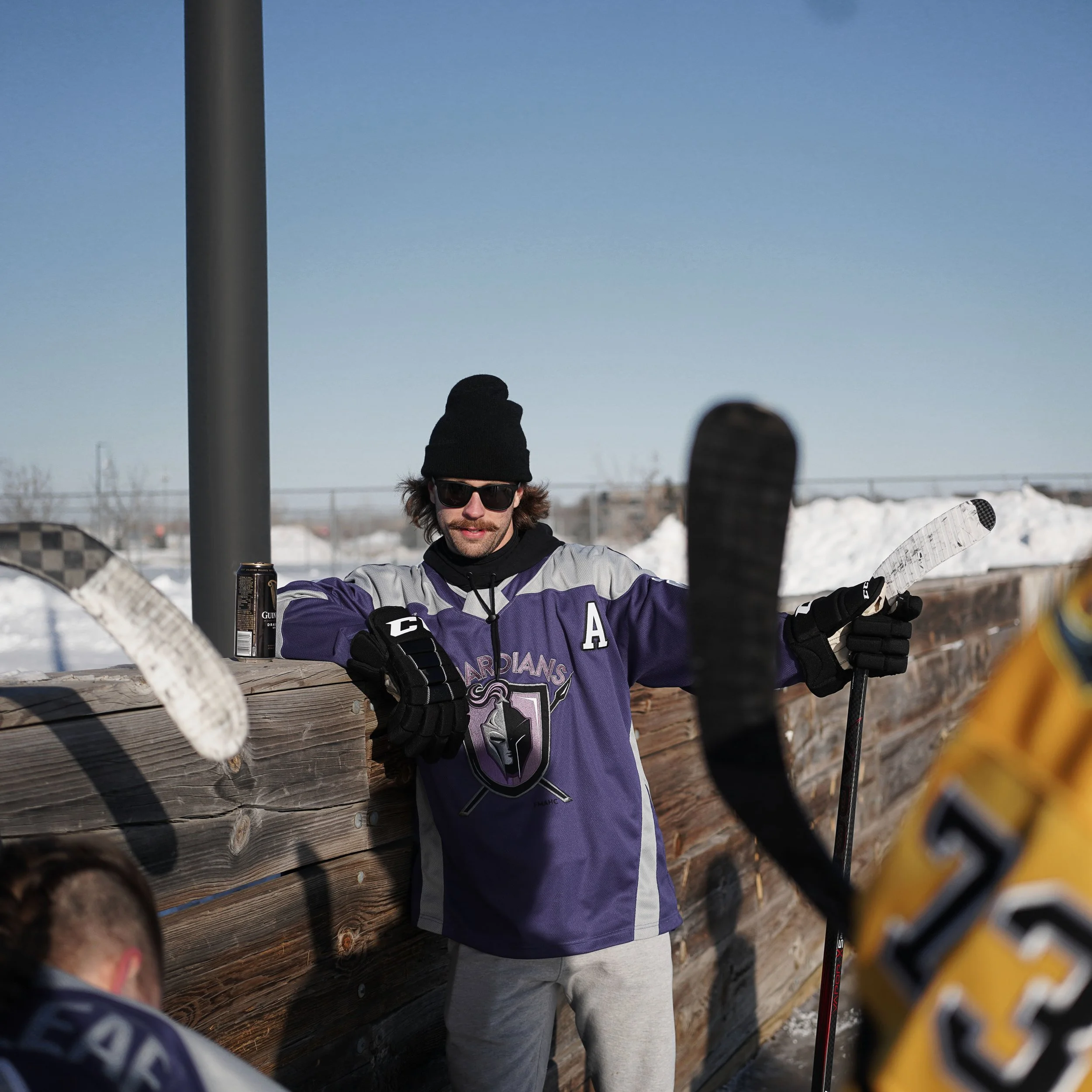 A man wearing a black beanie, sunglasses, and a purple hockey jersey with a logo and the letter 'A' on it leans against a wooden barrier holding a hockey stick. Drinking Guiness beer.