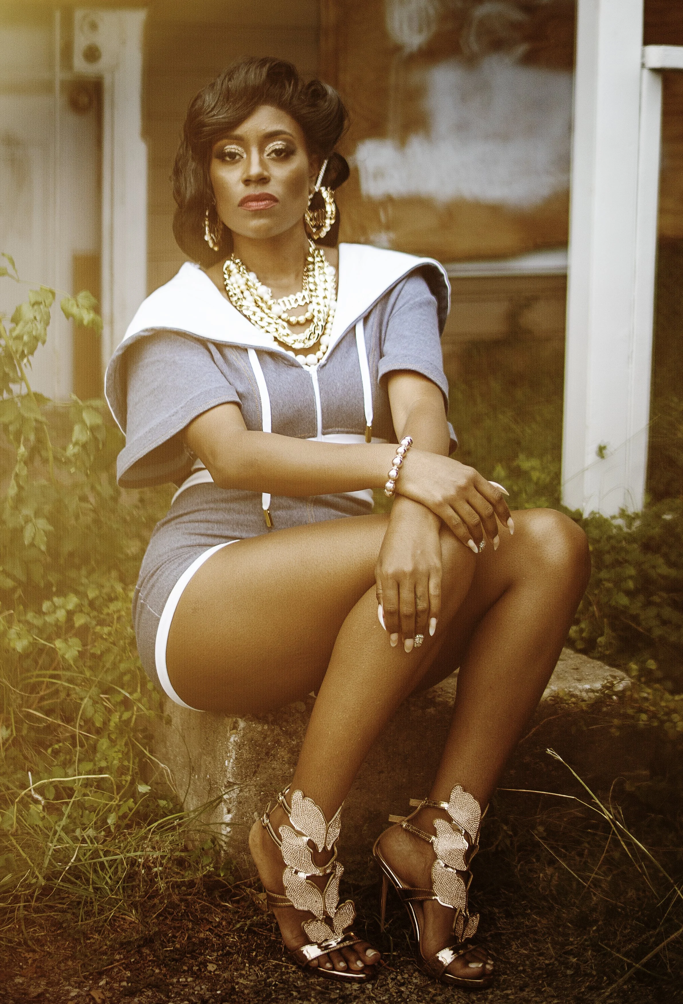 A woman with dark hair and gold jewelry sitting outdoors on a rock, wearing a gray outfit with a white hood, high heels with butterfly details, and makeup.