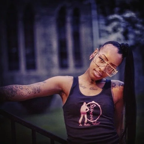 A woman with glasses and tattoos on her arms, wearing a tank top with a pink graphic, posing outdoors with a fence and trees in the background.