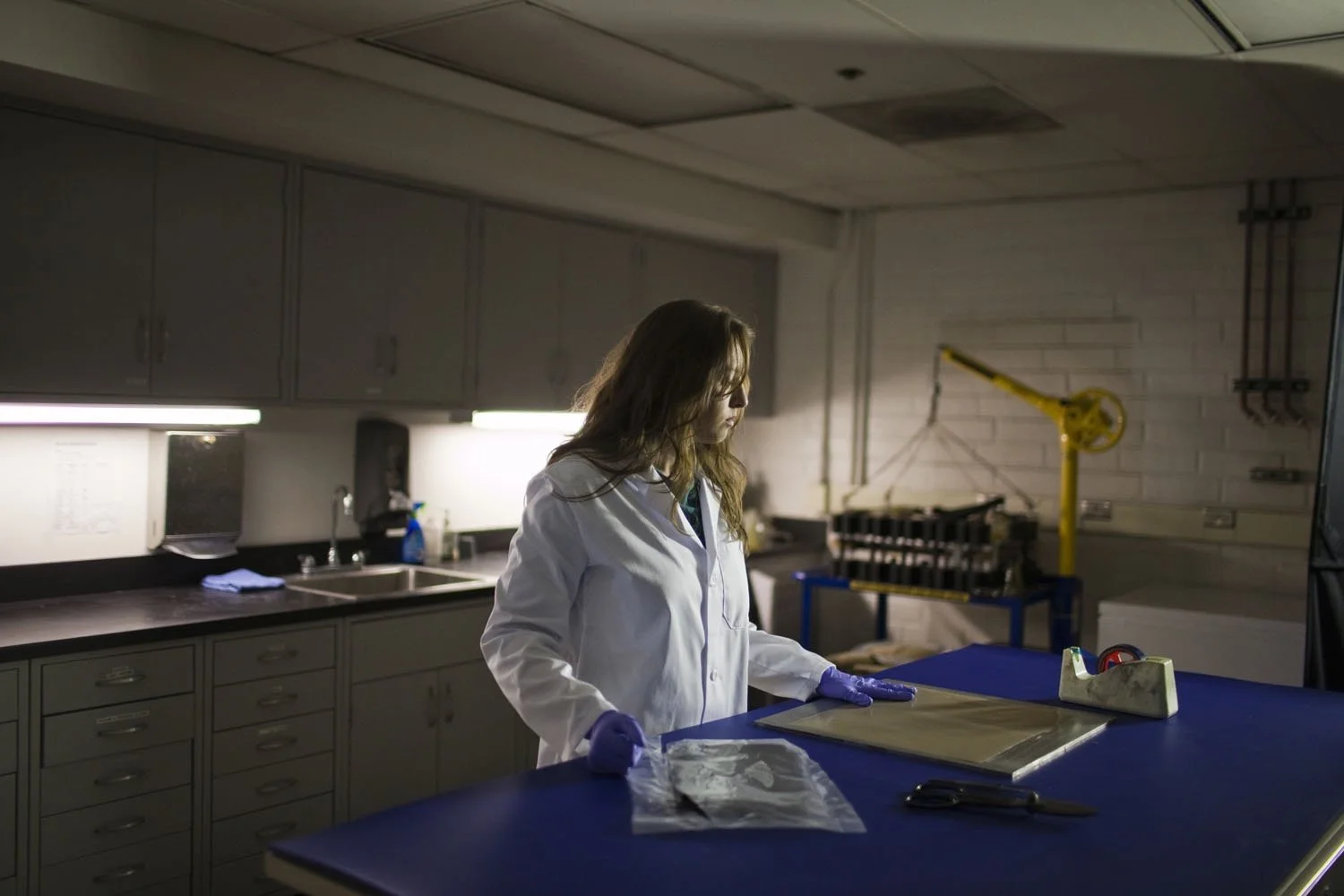 A woman in a white lab coat and purple gloves standing at a blue workstation in a laboratory, examining packages, with laboratory equipment and cabinets in the background.