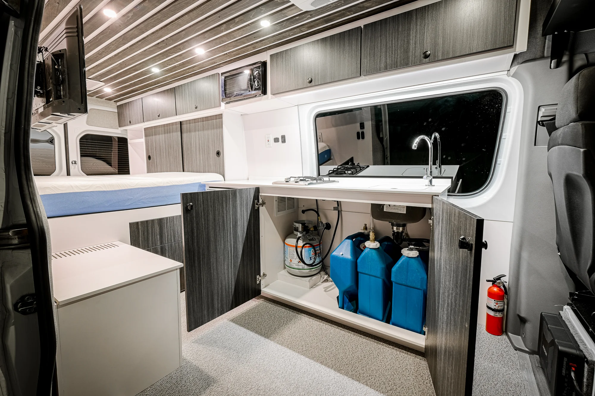Under-sink storage includes 2 7-gallon jerry cans for fresh water, one for gray/waste water, and a 20-lb propane bottle to feed the two-burner range as well as the rear-mounted shower system. 