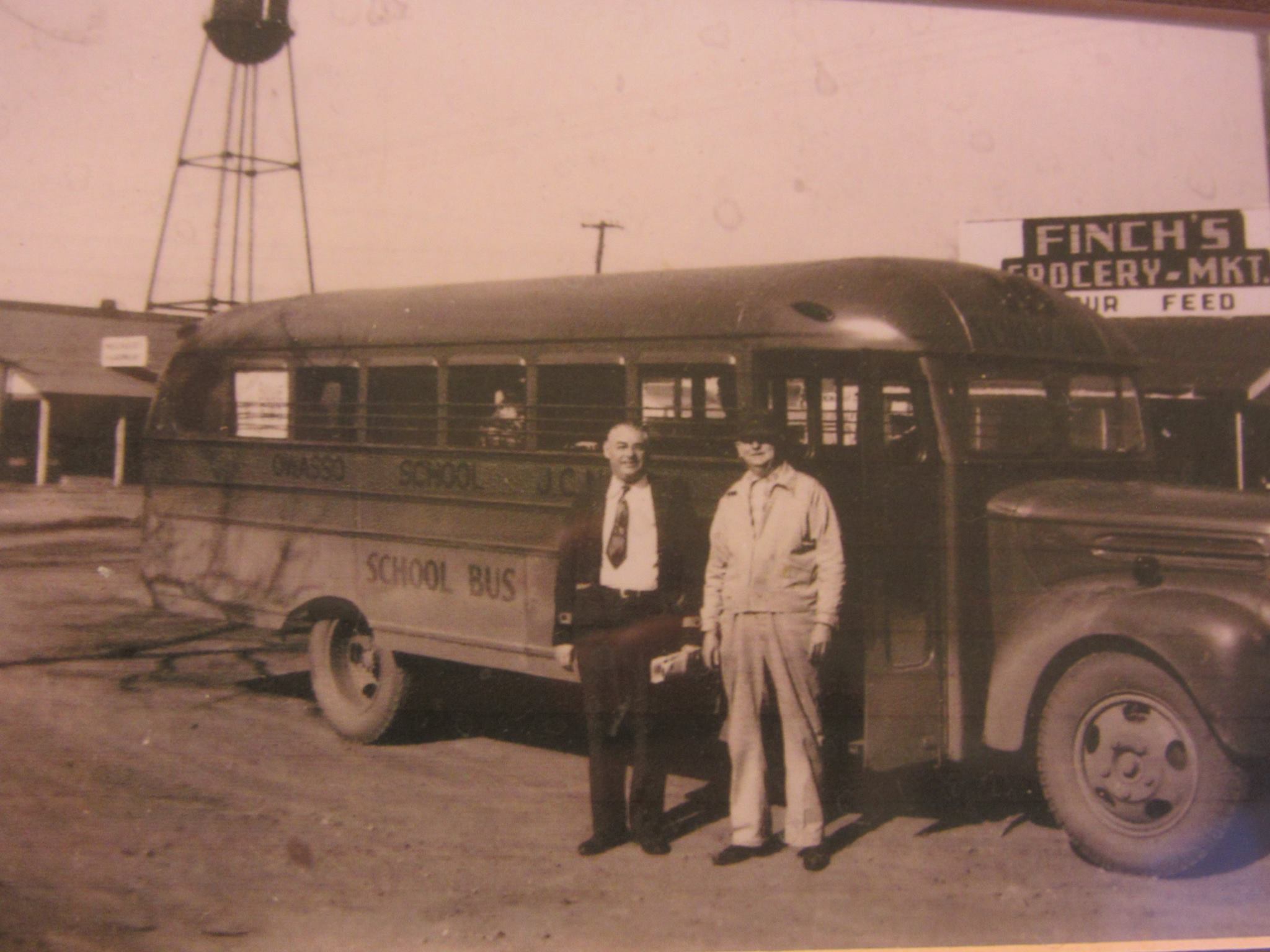 Early Owasso School Bus, Mr. Pinkerton, Superindent 1945-48, and Mr. Lewis Finch grocery owner. Finches Grocery was on East Broadway, Mounger Hardware is in the background on Main St. with the first water tower standing proud.