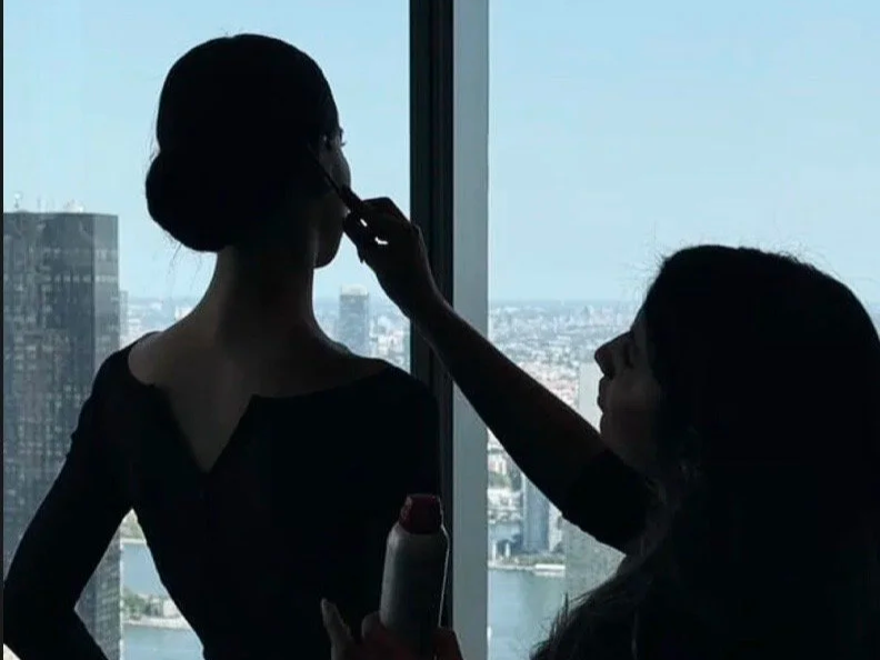 A woman getting her makeup done by a makeup artist in front of a large window with a city skyline view.
