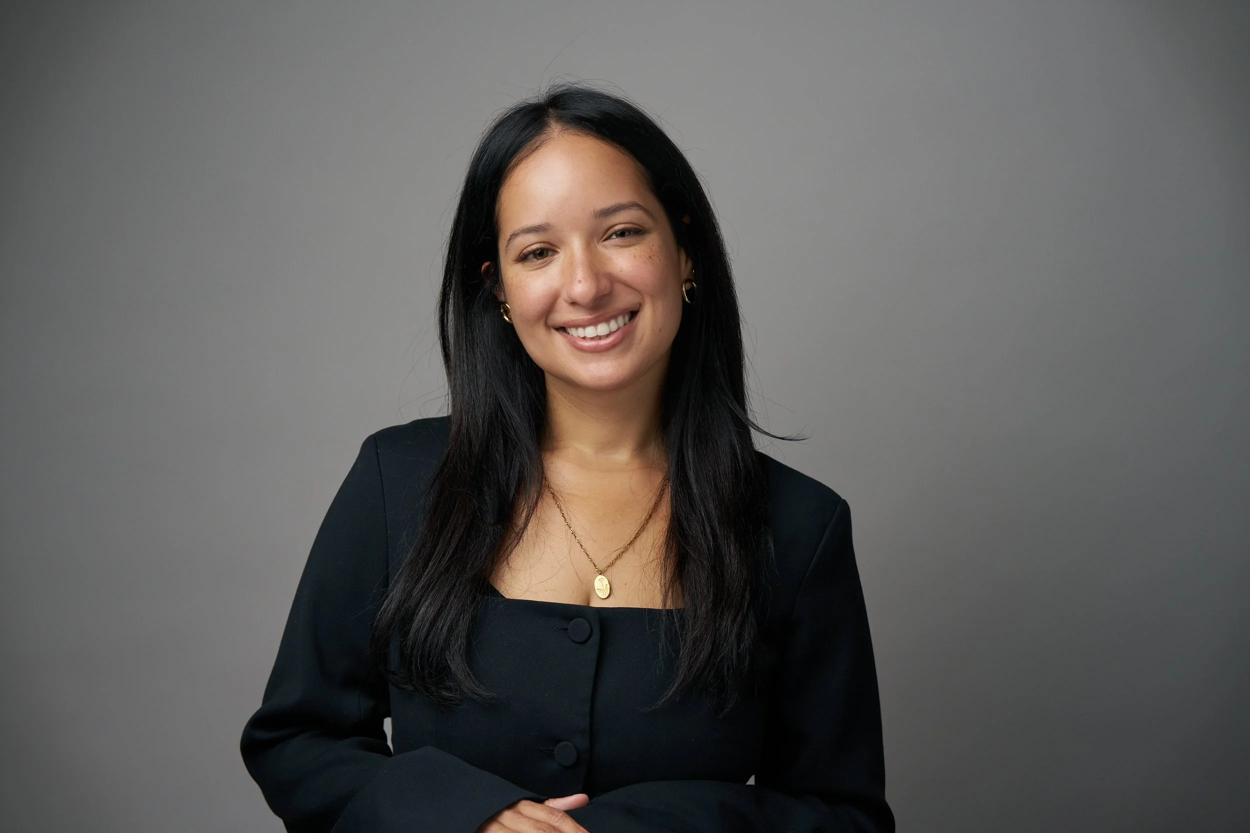 A young woman with long dark hair, smiling, wearing a black blazer and gold jewelry, standing against a neutral gray background.