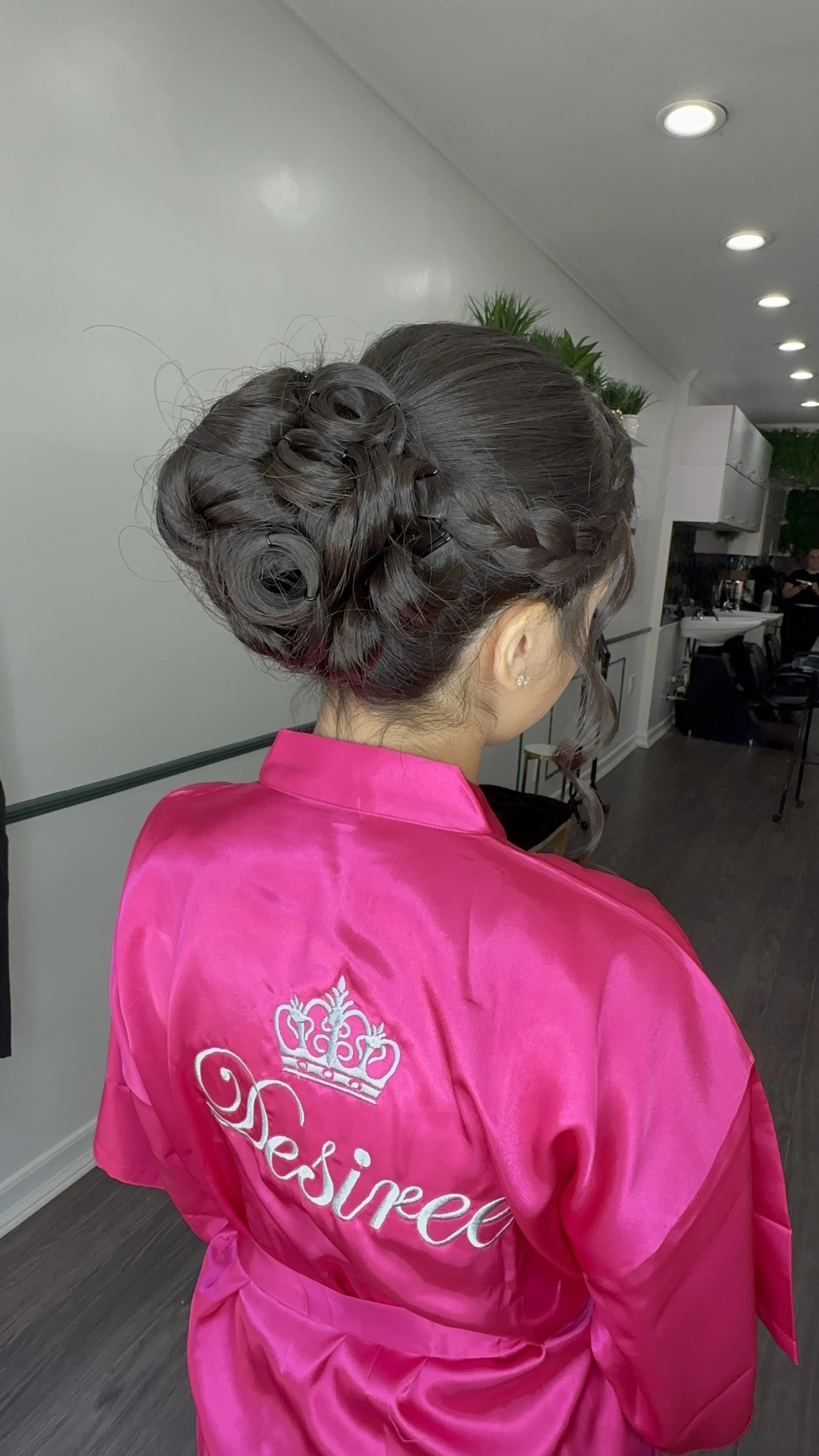 A woman with an elaborate updo hairstyle wearing a bright pink satin robe with the word 'Princess' embroidered on the back and a crown symbol.
