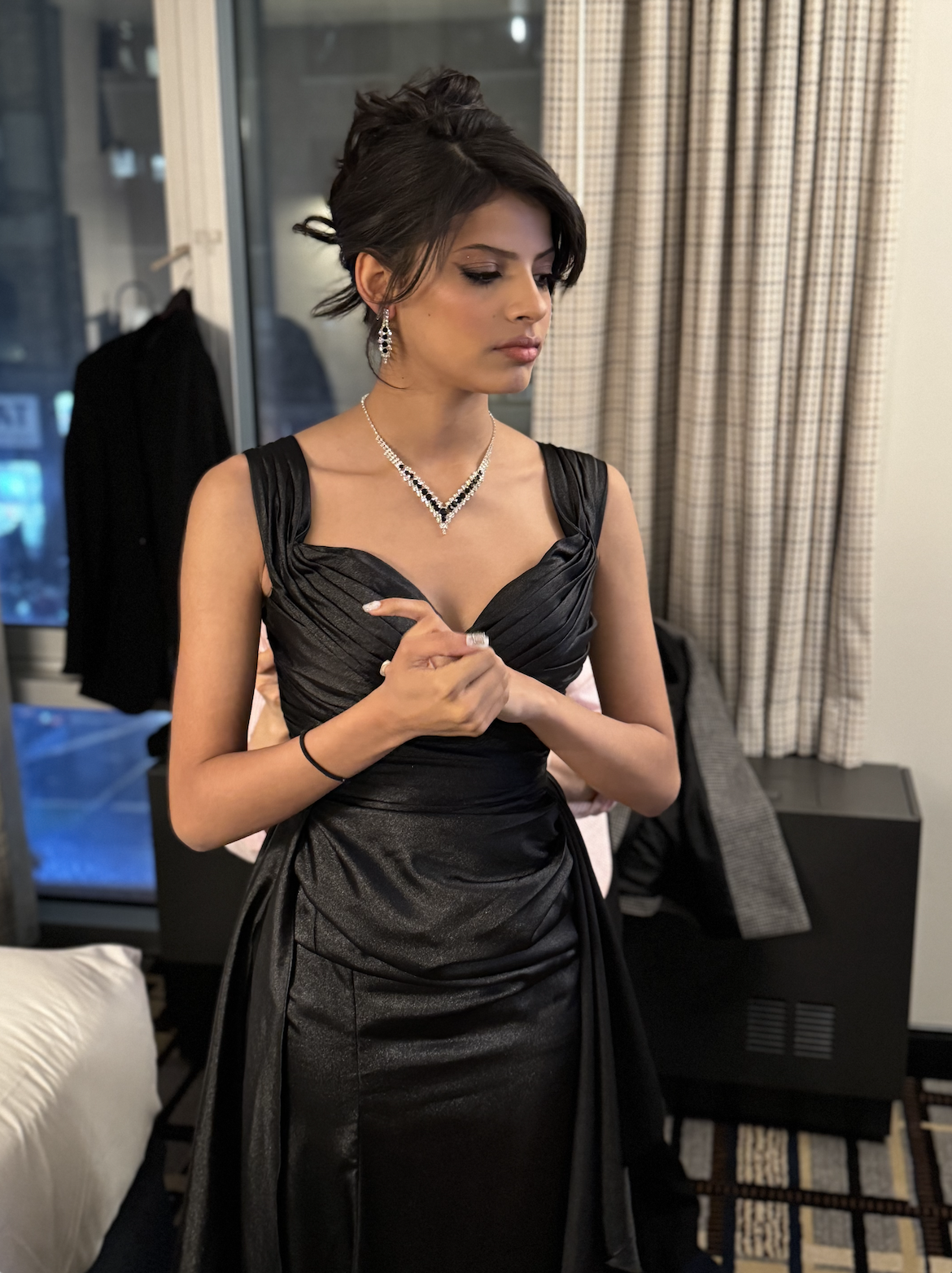 A young woman in a black evening gown with jewelry, standing indoors near a window and curtain, adjusting her ring, with a jacket hanging in the background.